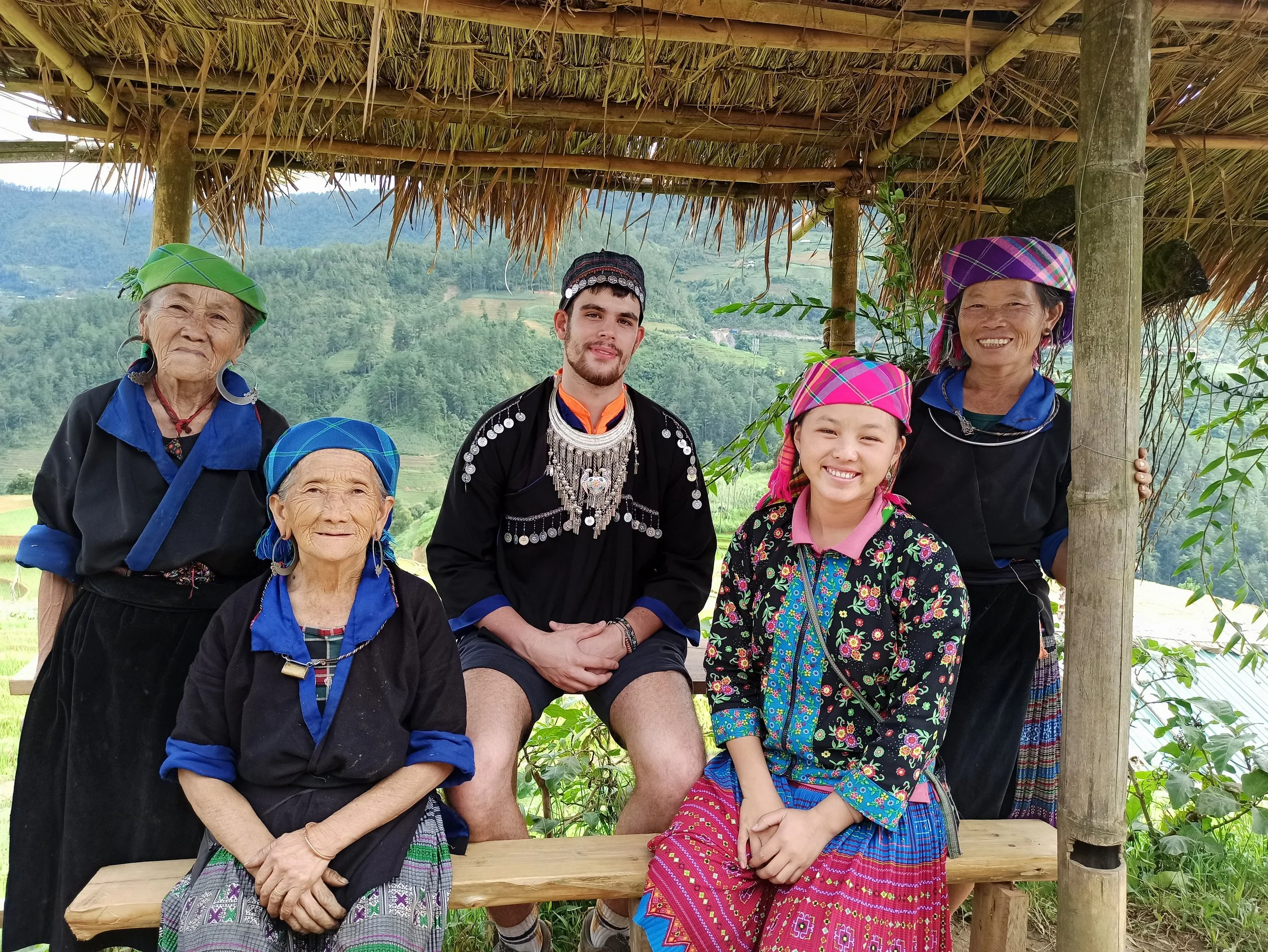 Groupe de personnes en vêtements traditionnels, deux femmes âgées et une jeune fille à gauche, un jeune homme au centre, et une femme plus âgée à droite, sous une structure avec un toit en feuilles de palmier, avec un paysage de montagnes en arrière-plan.