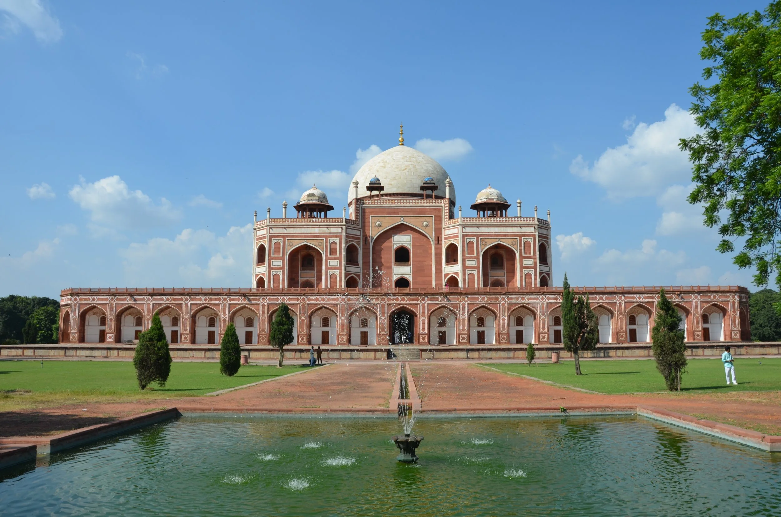 Le Taj Mahal, mausolée en marbre blanc à Agra, Inde, avec sa façade en pierre rouge, ses dômes et ses jardins verdoyants devant une allée d'eau et un ciel bleu.