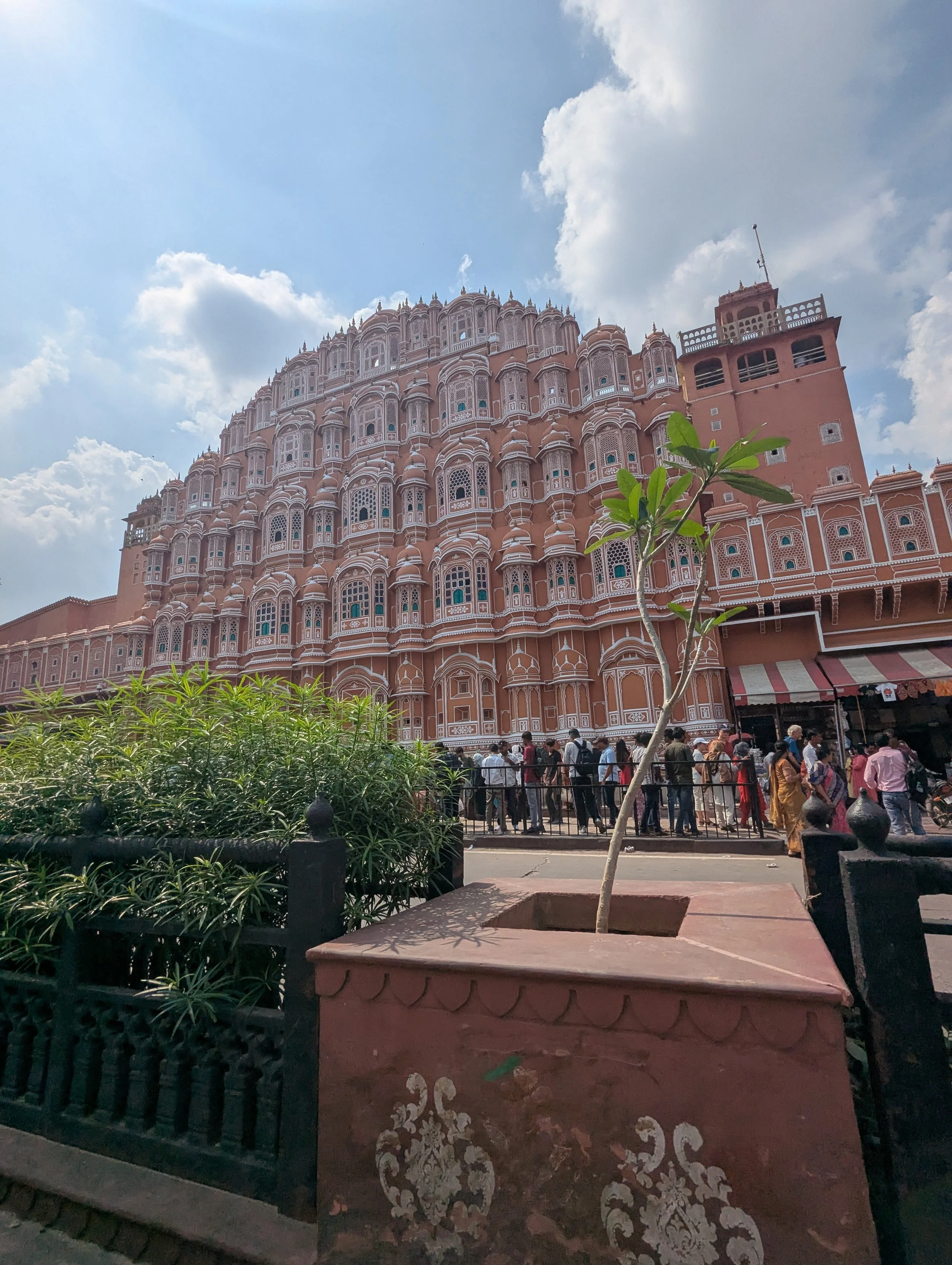 Un bâtiment rose très orné avec de nombreuses fenêtres, sous un ciel partiellement nuageux, avec des gens faisant la queue devant une entrée.