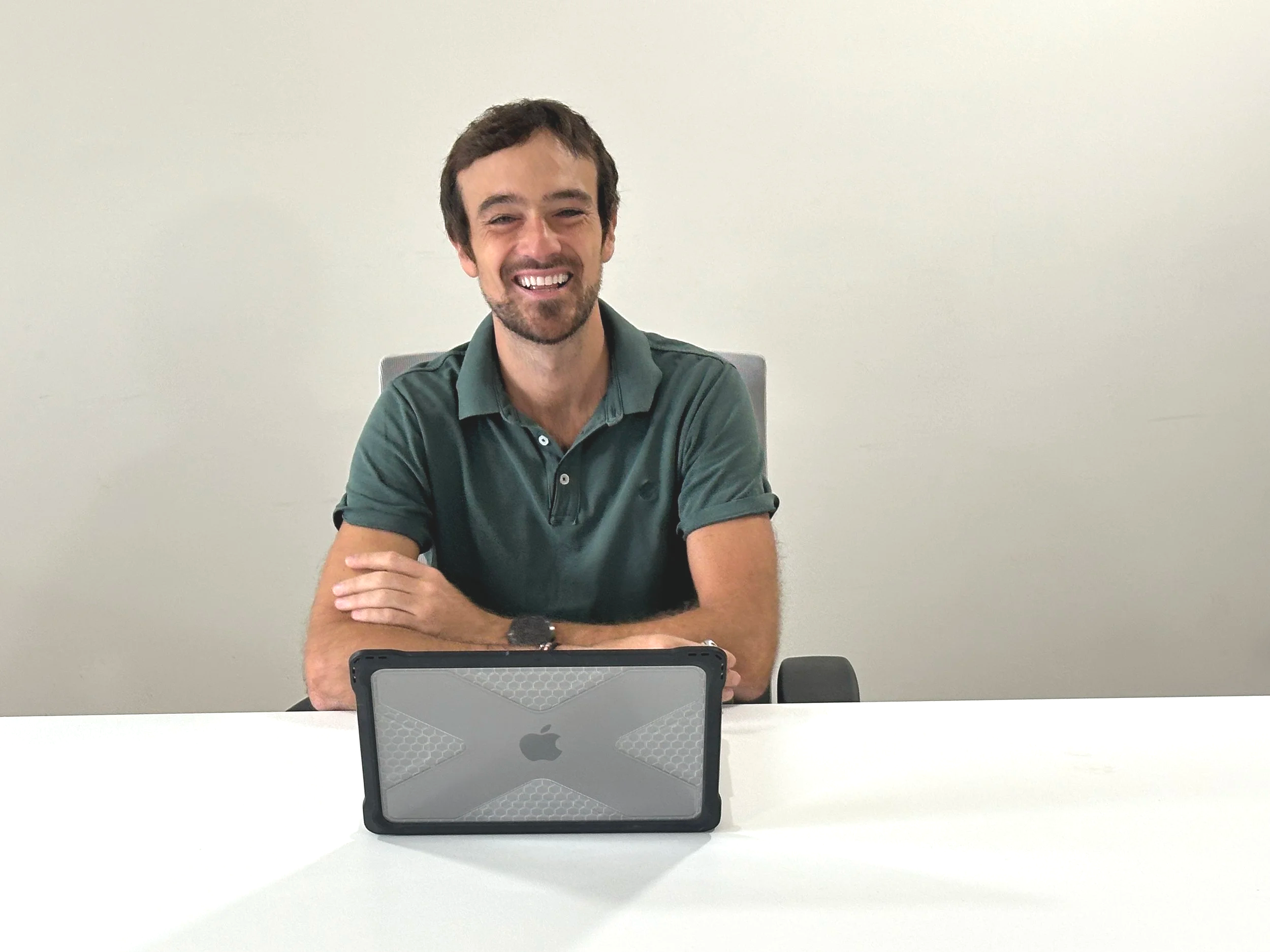Uomo con barba e capelli corti seduto a un tavolo bianco con un tablet Apple in primo piano, sorriso, maglietta verde, sfondo bianco.