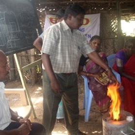 Man demonstrating use of an Anila stove, surrounded by onlookers