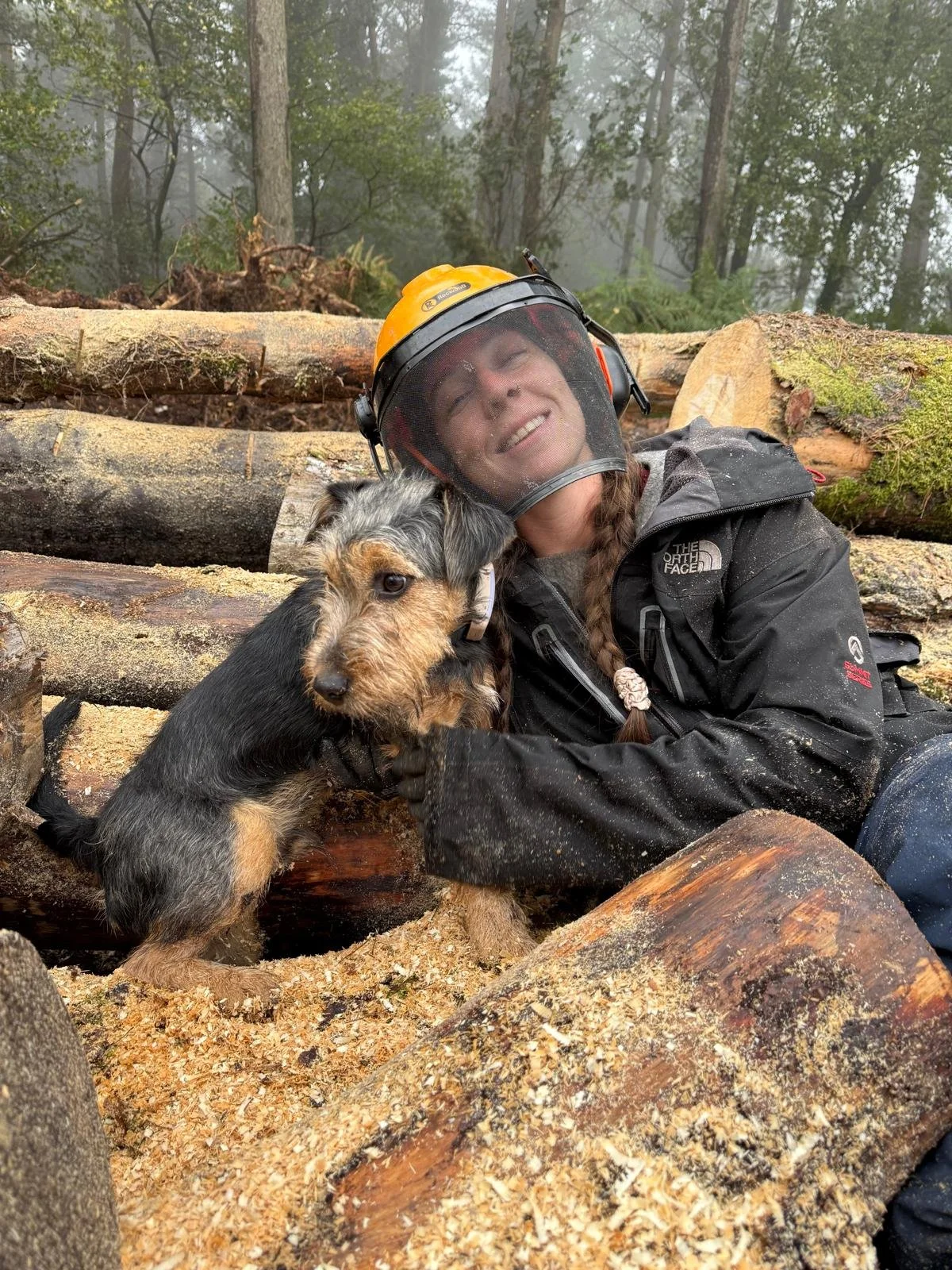 A person and a dog in a forest, with logs and sawdust around and the person wearing a helmet and a black outdoor jacket, smiling as they pose together.