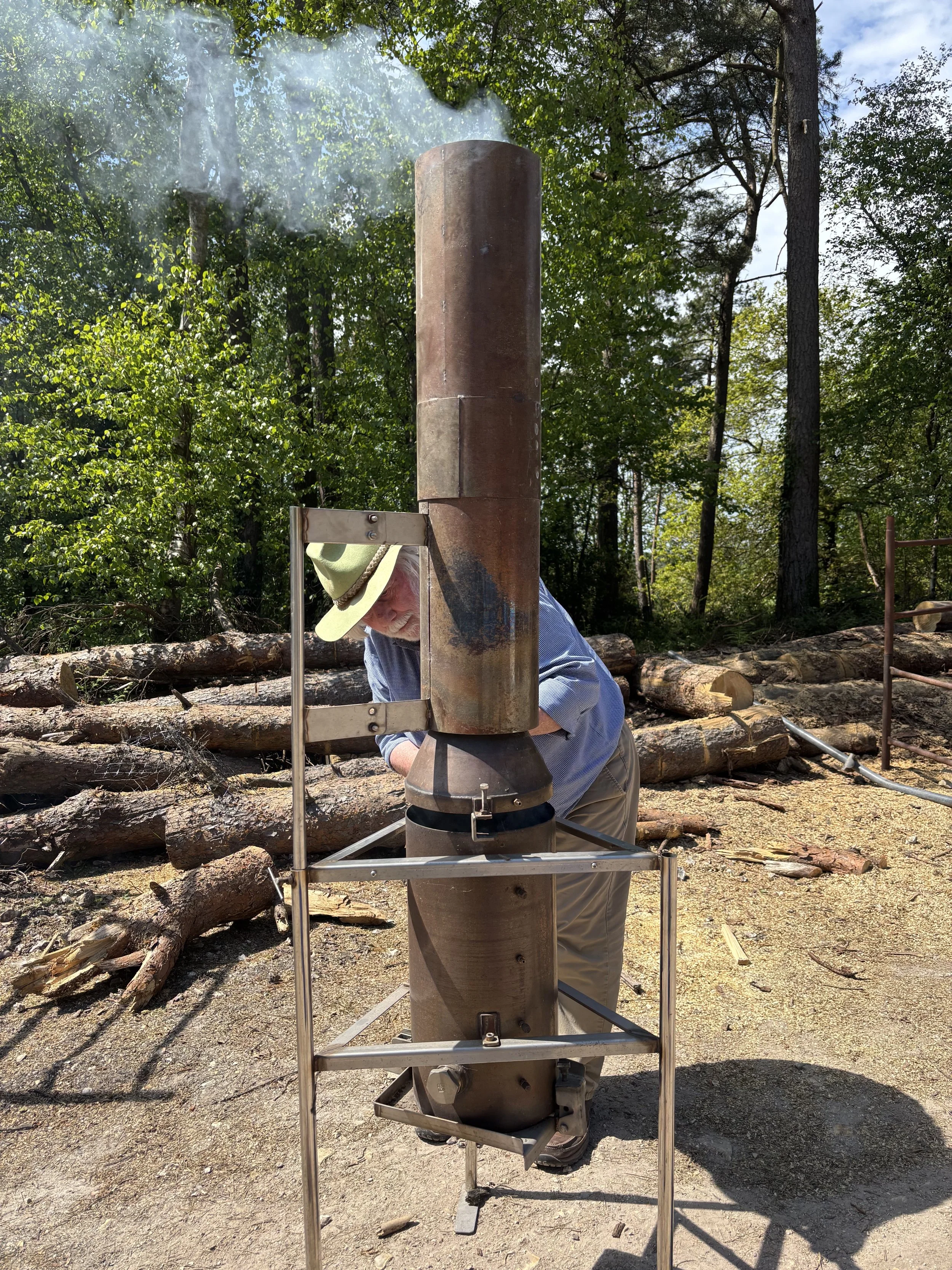 An older man wearing a light-colored hat and blue shirt is tending to a demonstration biochar kiln in a forested area, with logs in the background.