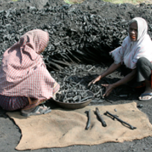 Two women sitting on the ground near a pile of biochar, using tools for biochar production.