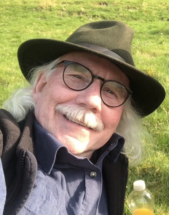 A smiling elderly man with white hair, mustache, and glasses, wearing a wide-brimmed hat and a dark jacket, is outdoors in a grassy field.