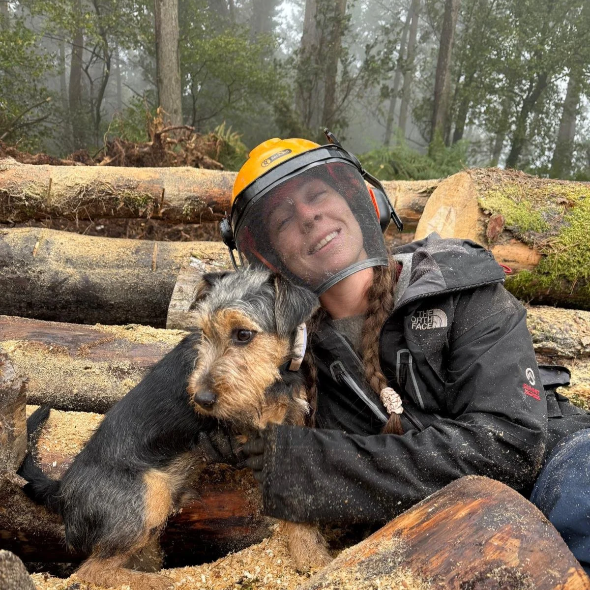 A woman in outdoor chainsaw gear, wearing a chainsaw helmet and a black work jacket, smiling while lying on logs in a forest, cuddling a small, scruffy dog with black and gray fur.