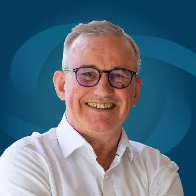 Richard Ord with gray hair, wearing glasses and a white collared shirt, standing against a blue geometric background.