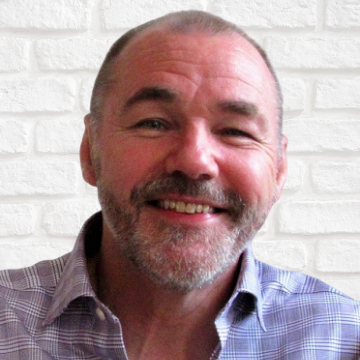 David Stewart with a beard and short hair, wearing a checkered shirt, standing in front of a white brick wall.