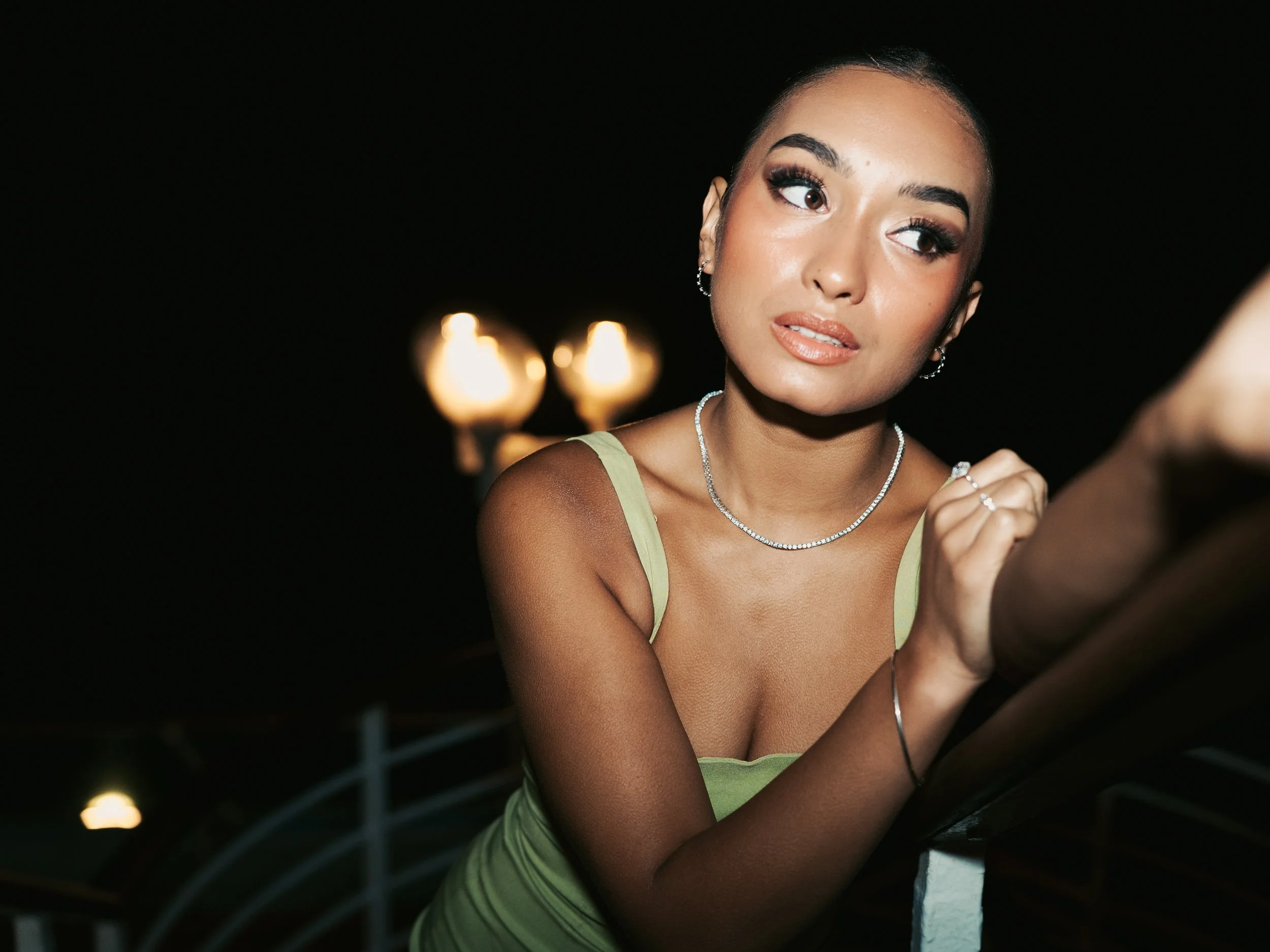 A woman with dark hair styled in a bun, wearing a light green spaghetti strap dress, silver jewelry, and makeup, looking to her left, with a dark background and blurred lights behind her.
