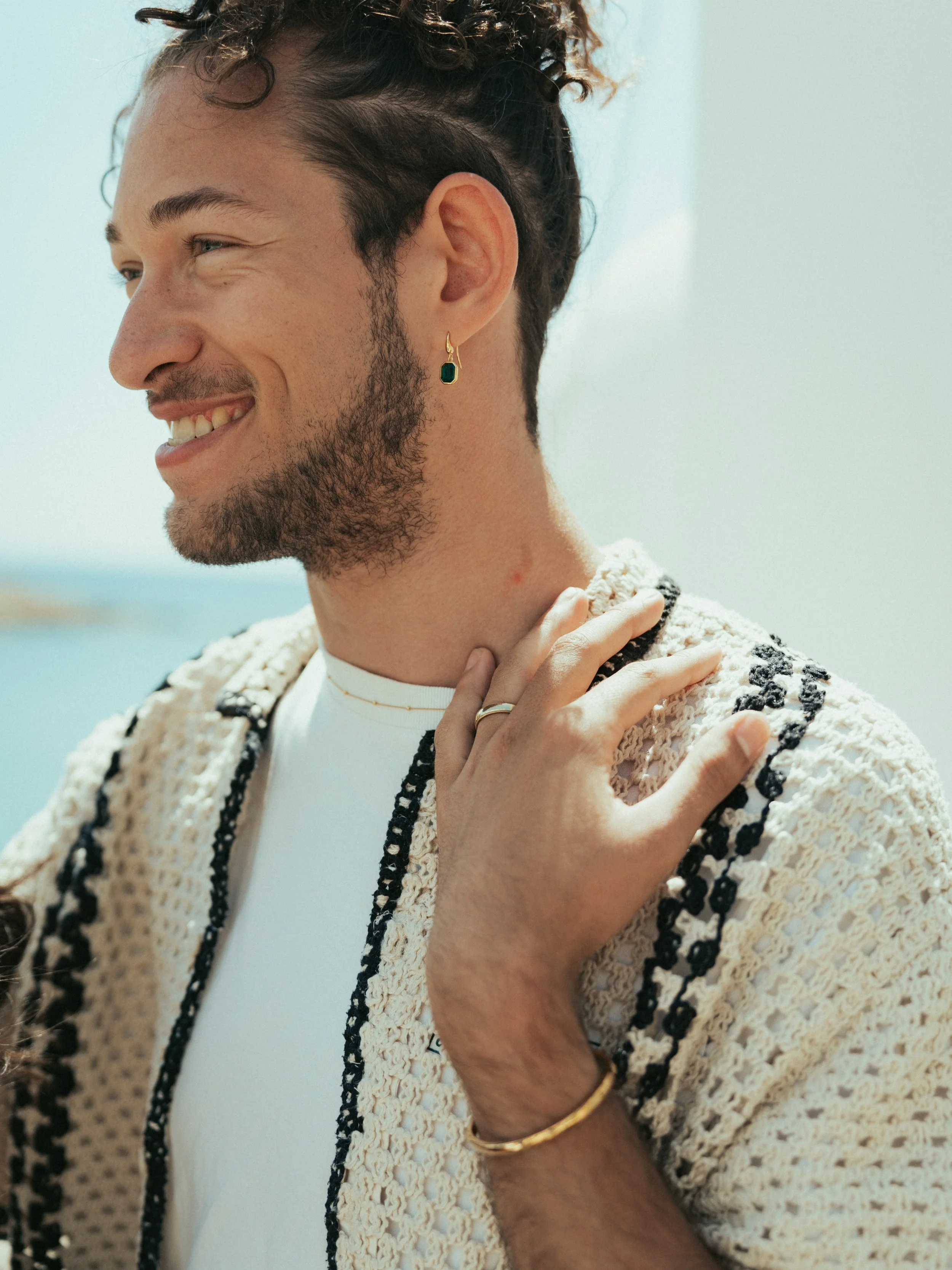 Close-up of a smiling man outdoors, wearing jewelry including an earring, ring, bracelet, and a thin necklace, with a blurred background of water and sky.