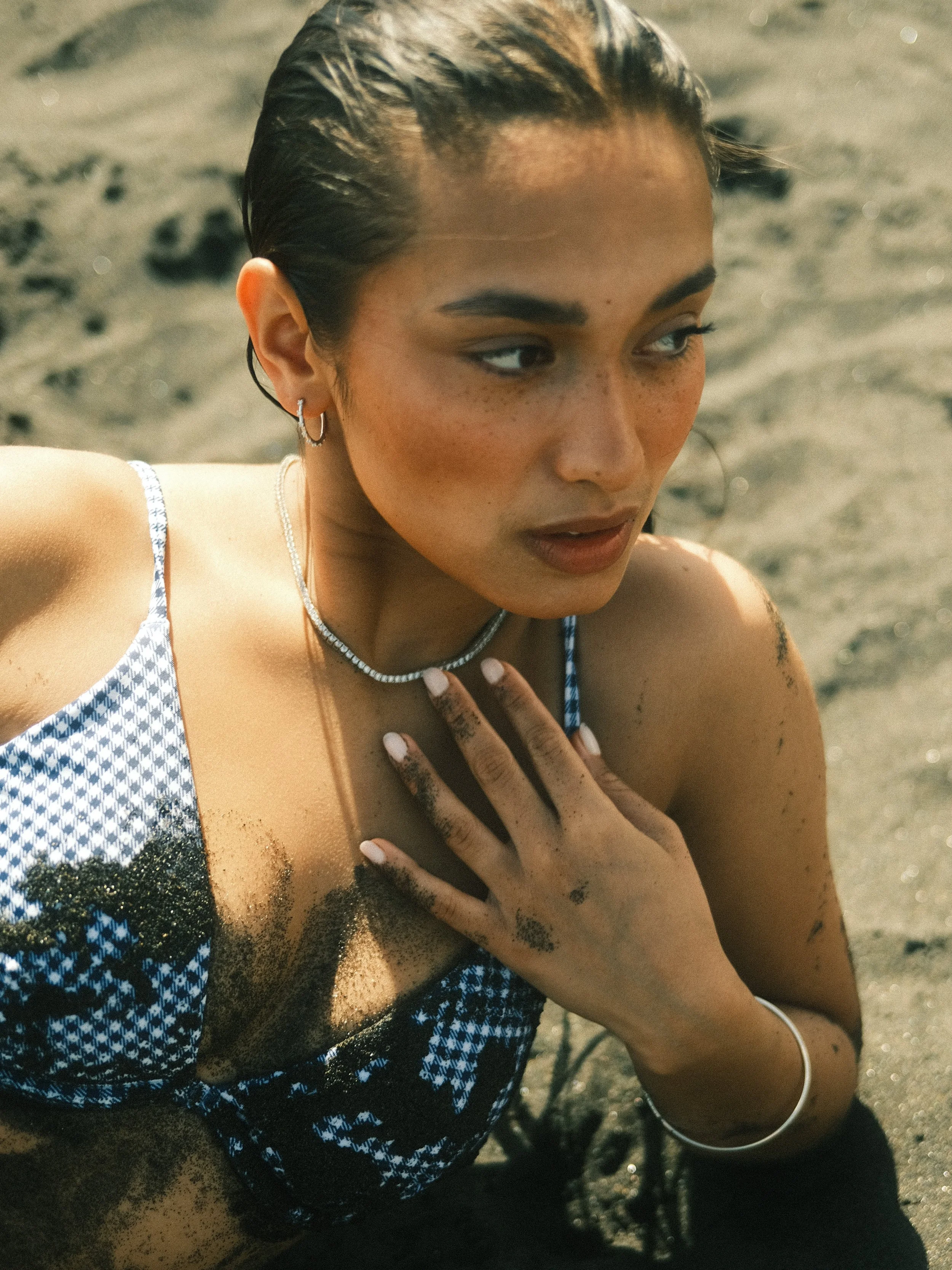 A woman with wet hair and fair skin lying on sandy beach, wearing a blue and white patterned swimsuit, with sand on her skin, adorned with jewelry, looking thoughtfully to the side.