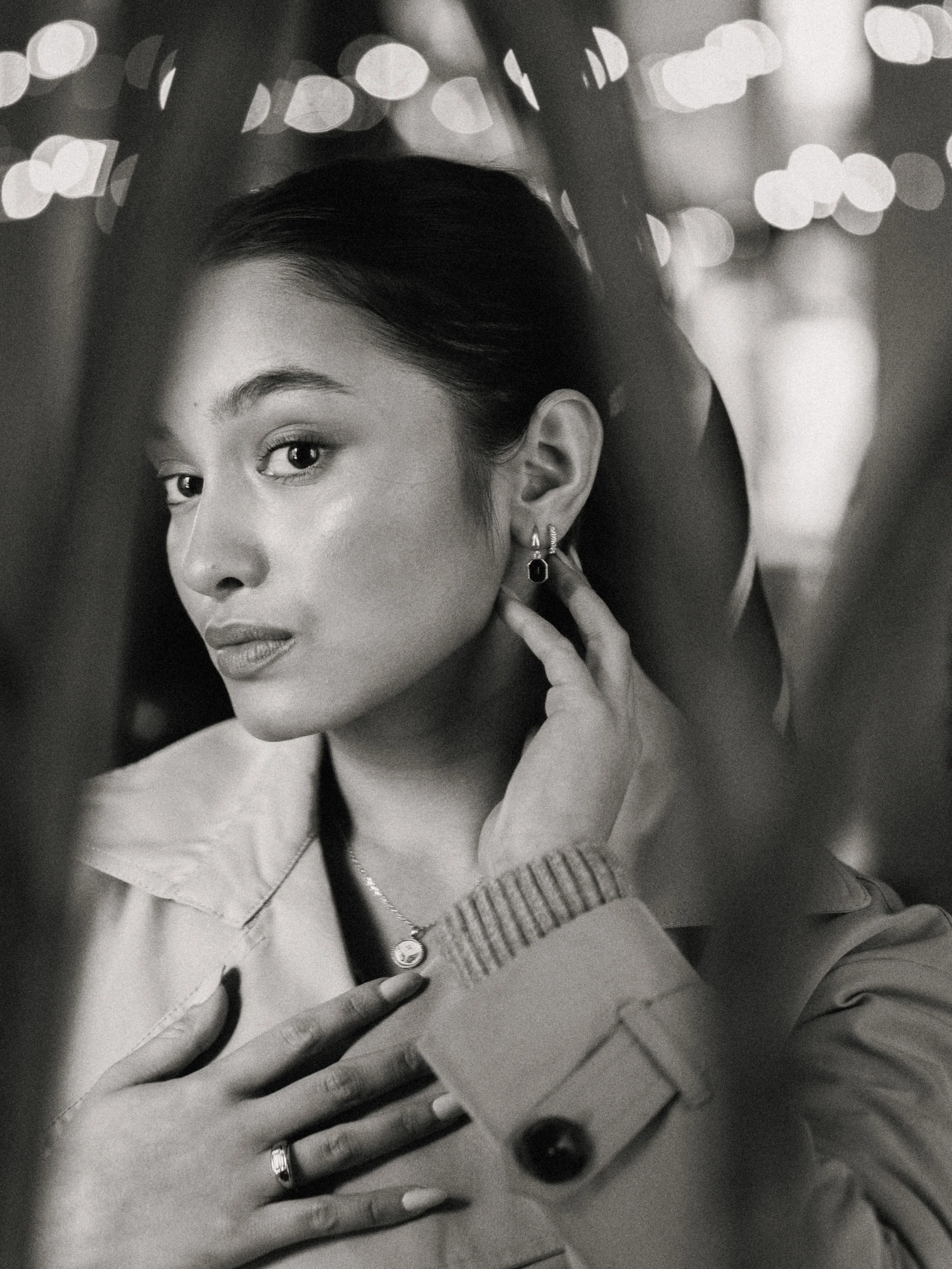 A black and white portrait of a young woman touching her earrings with her right hand and wearing a necklace and rings, with a blurred background of lights.