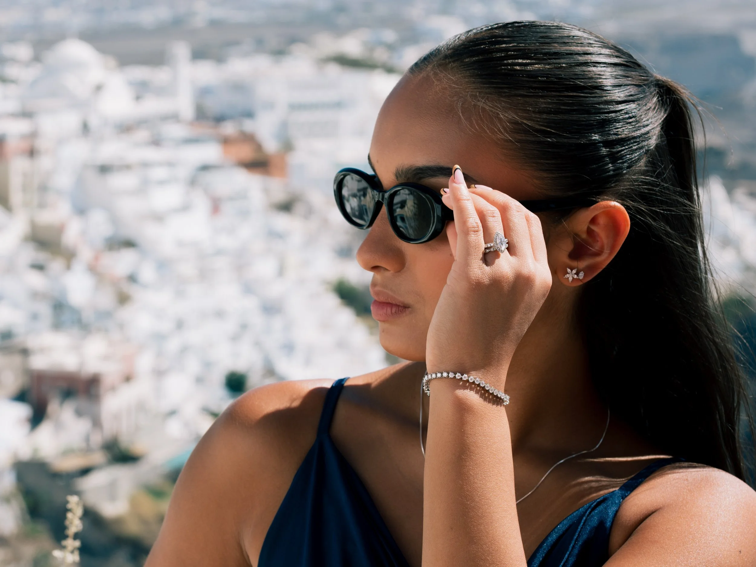 A woman with long dark hair wearing black sunglasses, diamond jewelry, and a dark blue dress, adjusting her sunglasses against a blurry coastal cityscape background.
