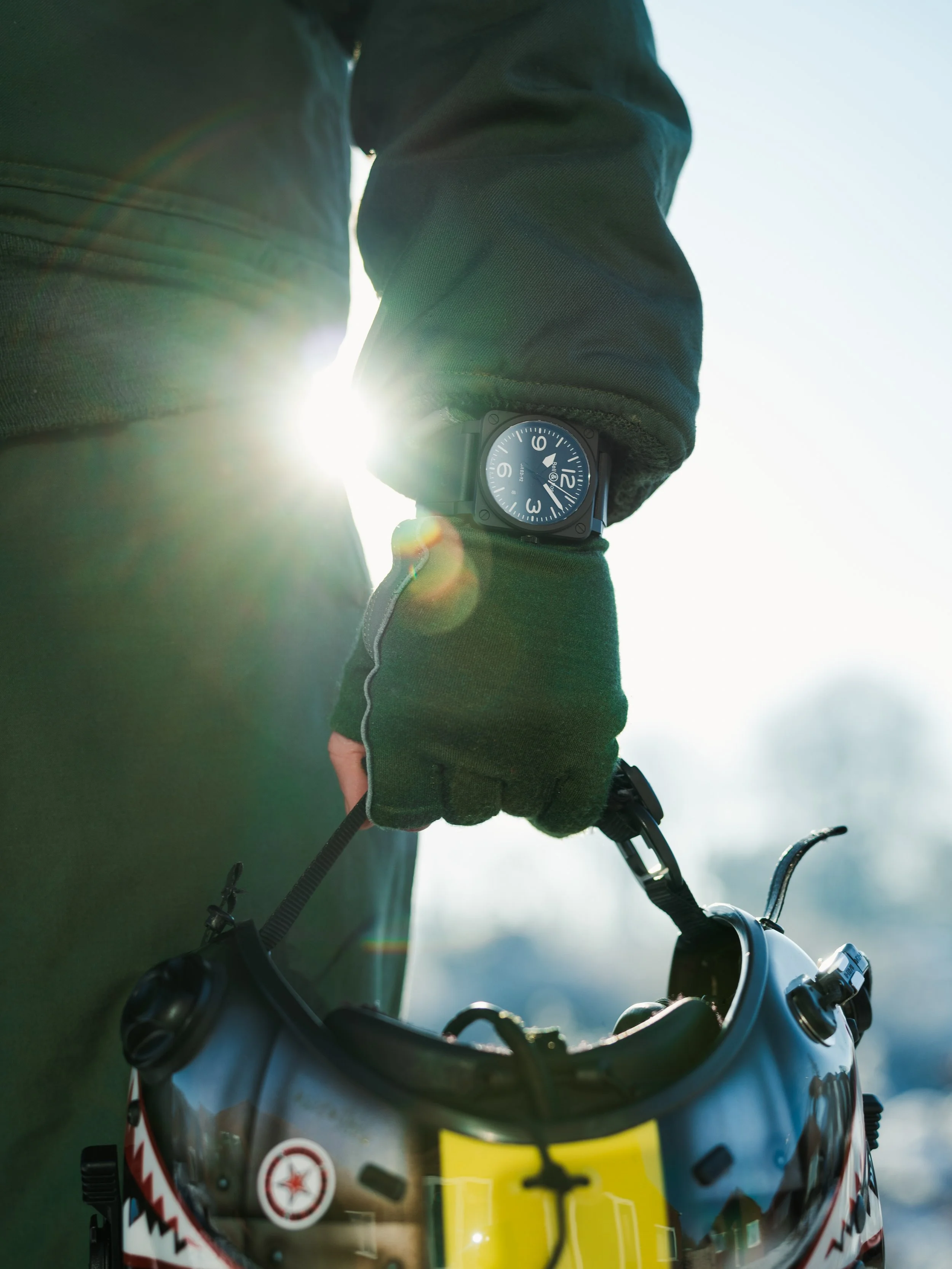 A person wearing a green jacket and gloves holding a helmet with goggles, standing outdoors in sunlight.