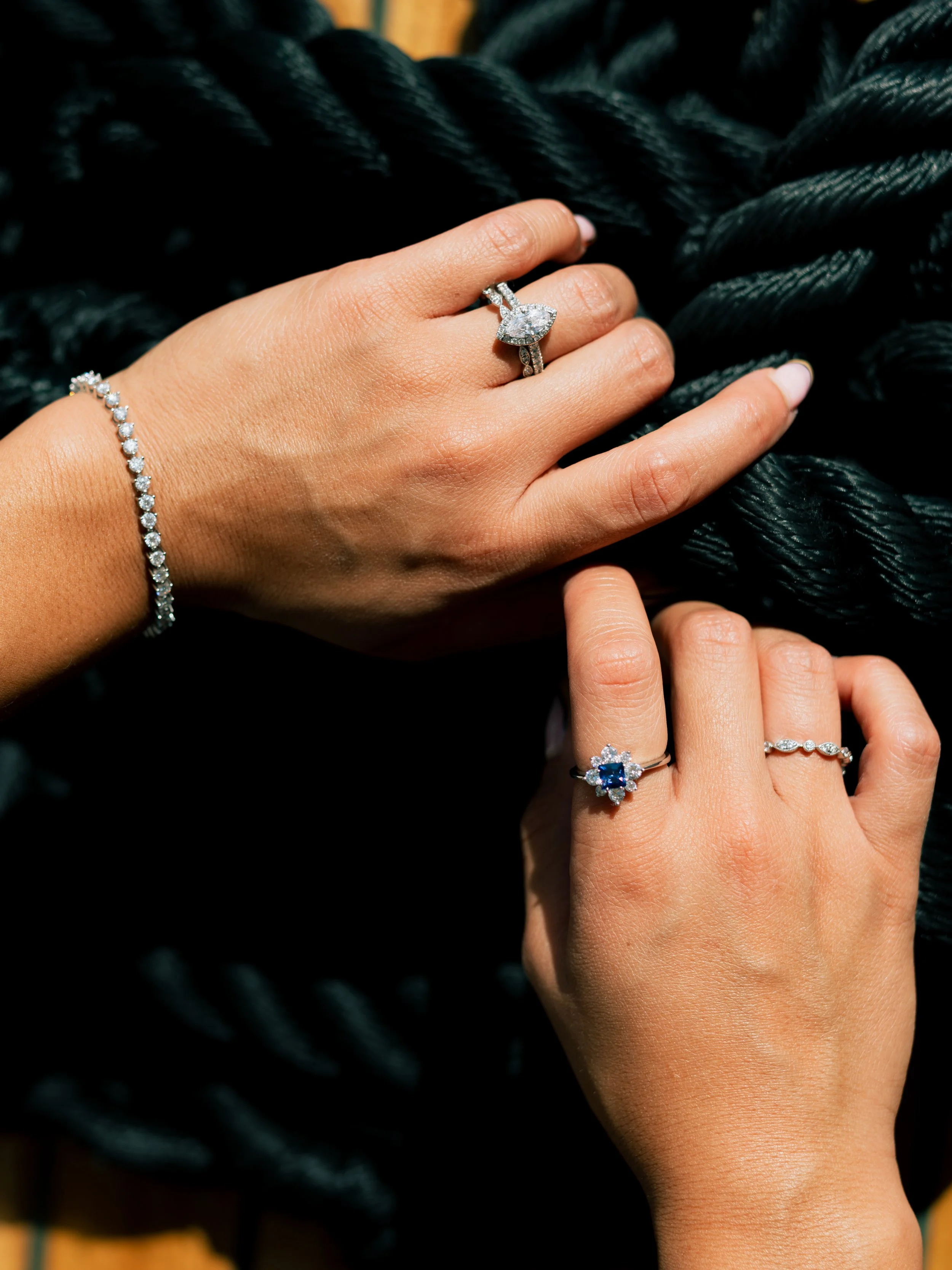 Close-up of two hands with multiple diamond rings and bracelets resting on a black textured surface.