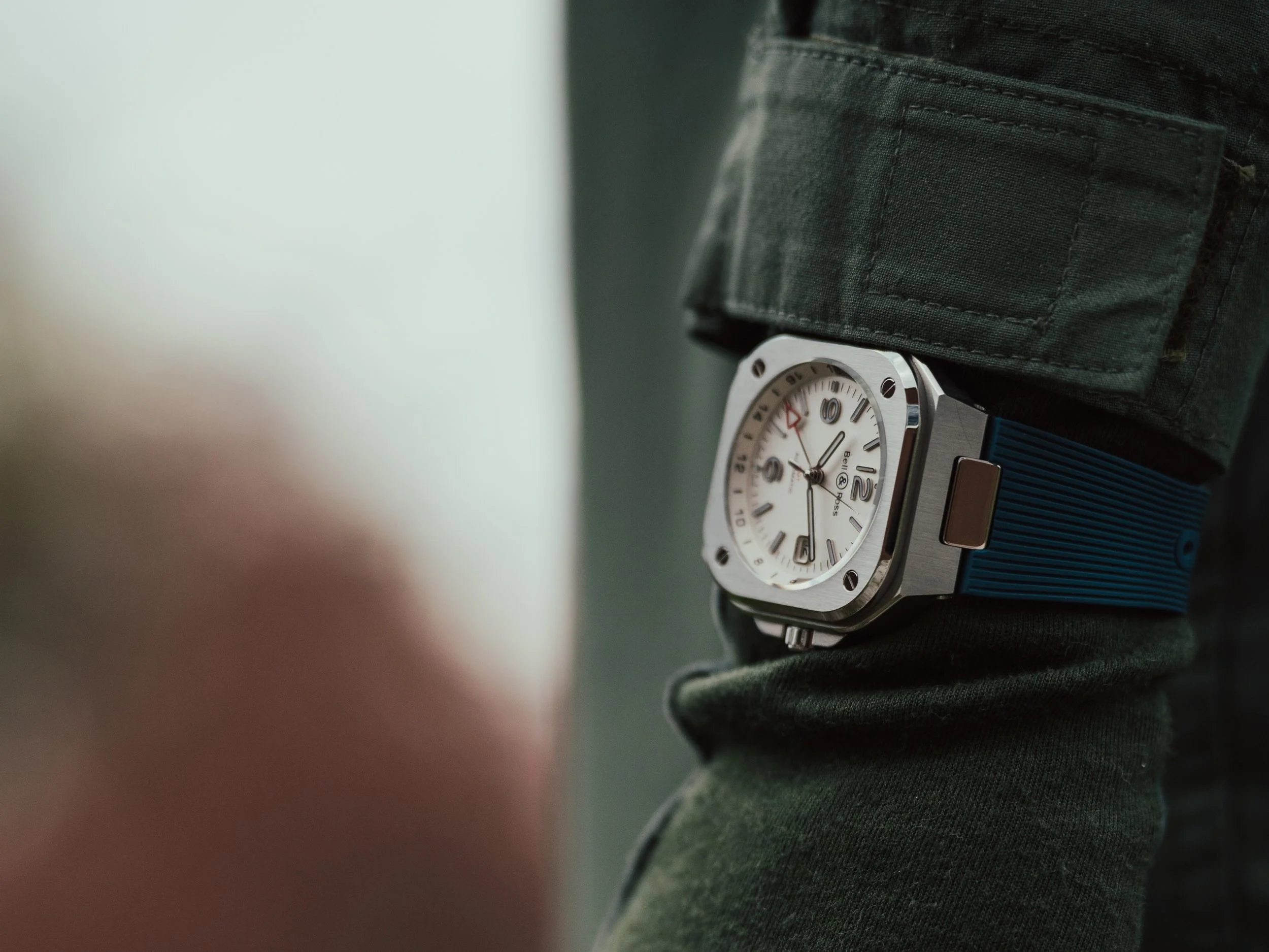 A close-up of a person's wrist showing a square-faced watch with a white dial, black numerals, and a blue strap, worn over a dark green jacket sleeve.