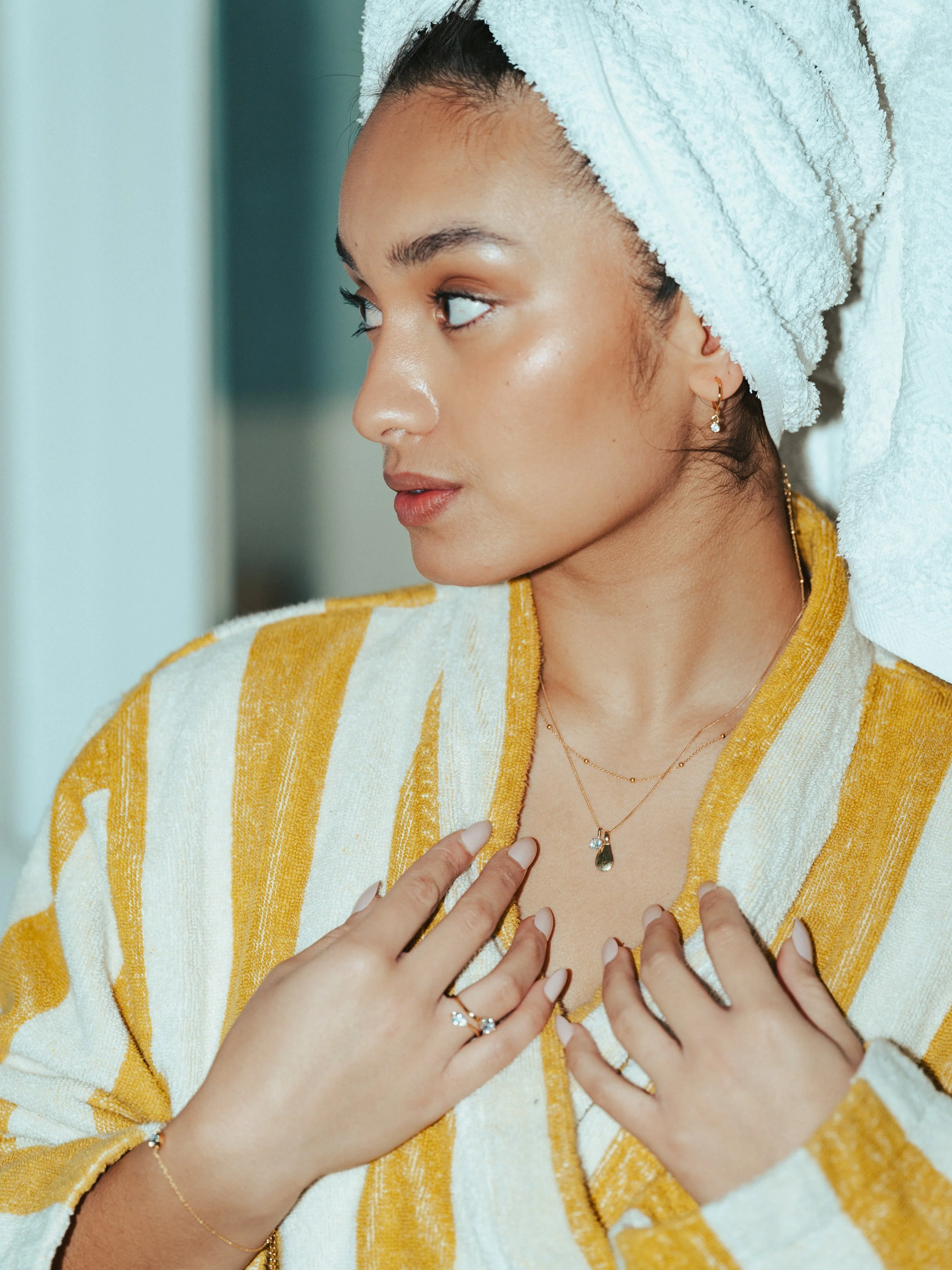 A woman wrapped in a yellow and white striped towel with a white towel on her head, wearing jewelry, looking to the side.