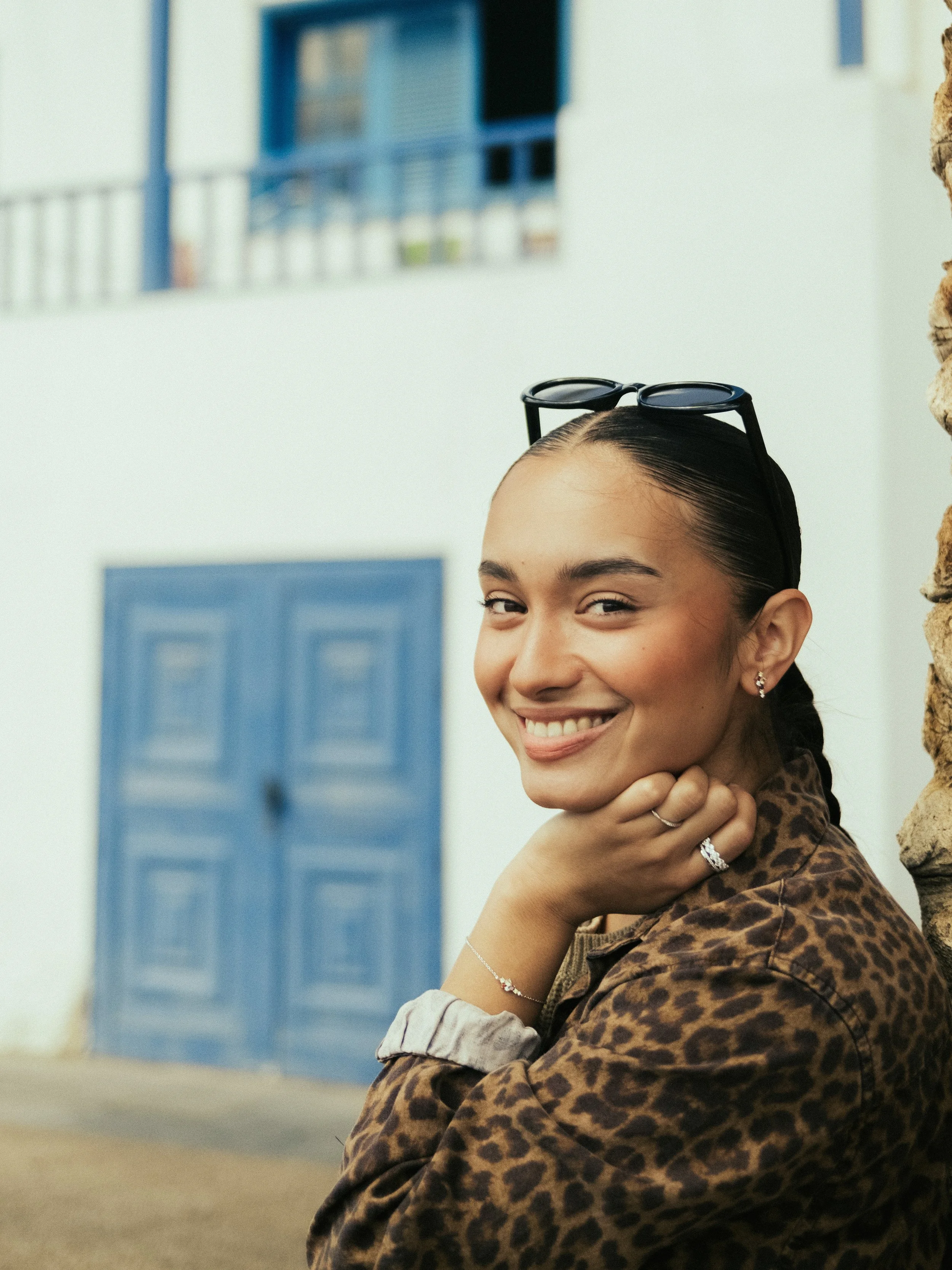 Smiling woman with glasses on her head, jewelry, and a leopard print jacket, sitting outdoors near a stone wall with a white building and blue window shutters in the background.