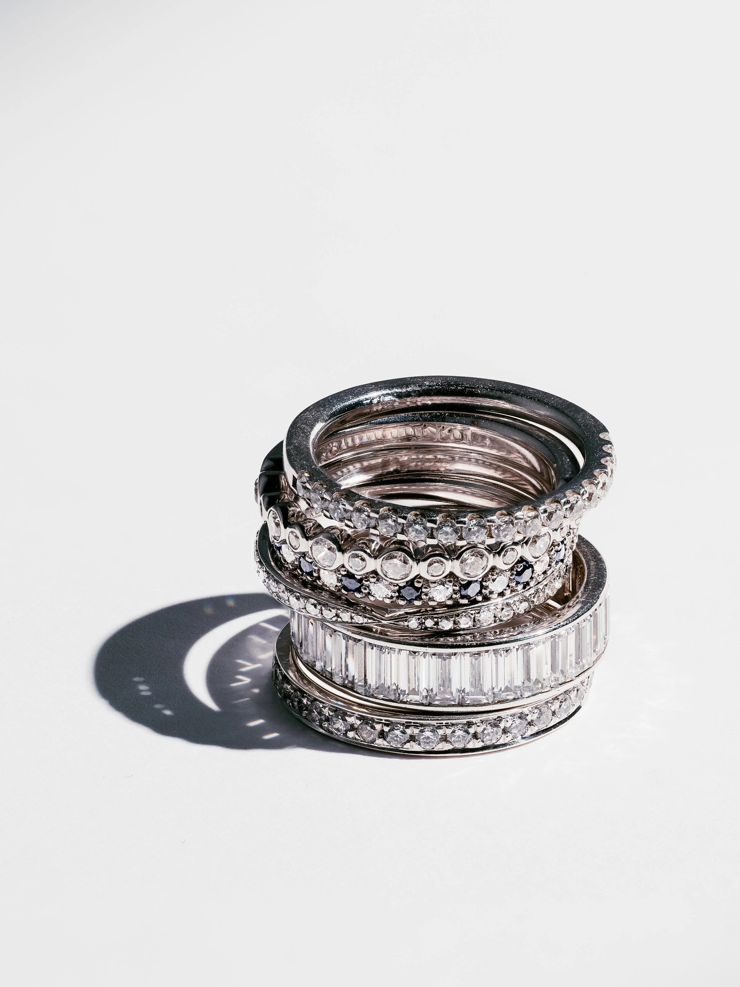 Stack of silver rings and bangles decorated with numerous clear and black gemstones on a white background, casting a shadow.