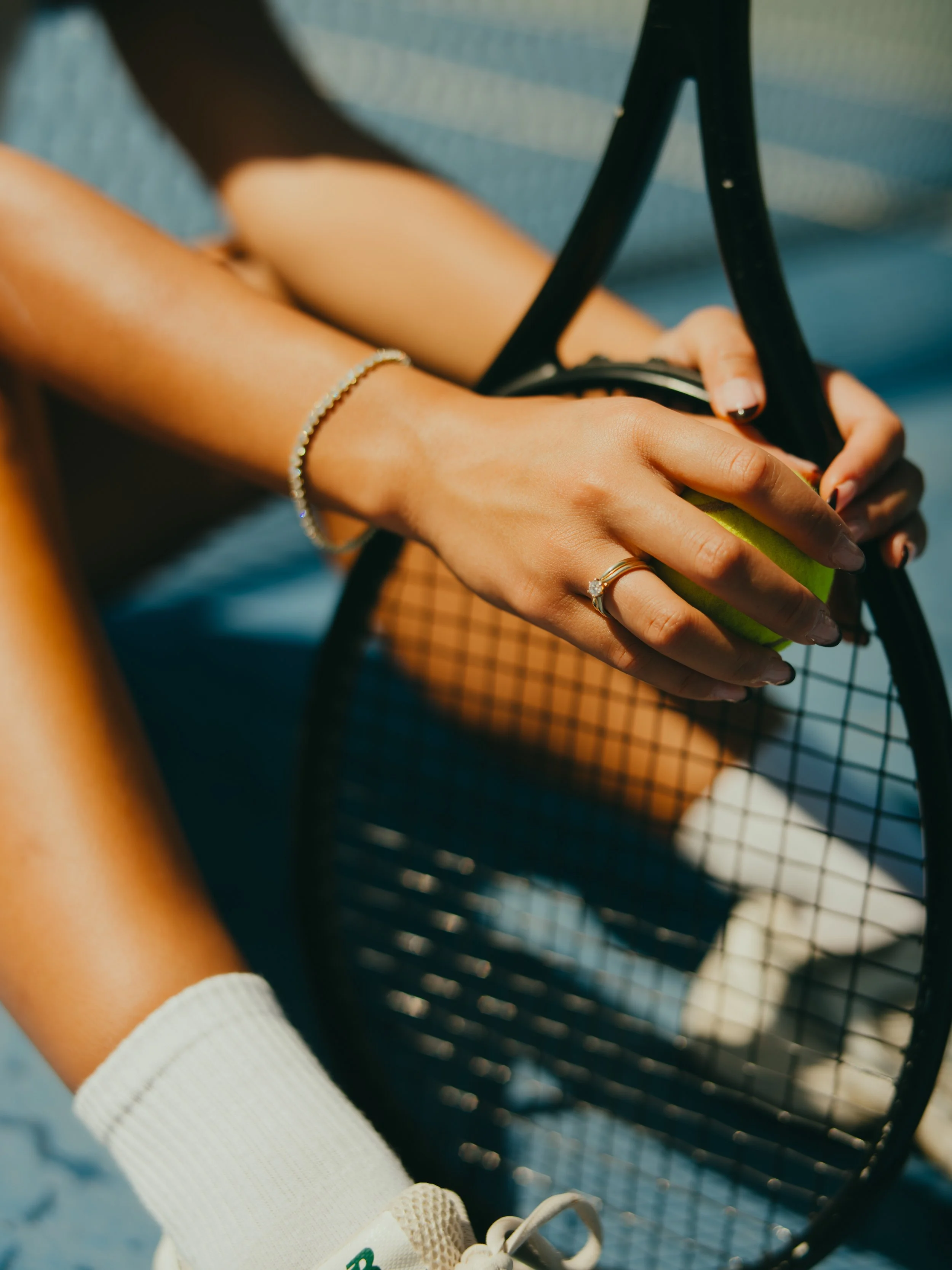 A person's hand with jewelry, holding a tennis racket and a green tennis ball, sitting on a blue tennis court, wearing white tennis shoes and black nail polish.