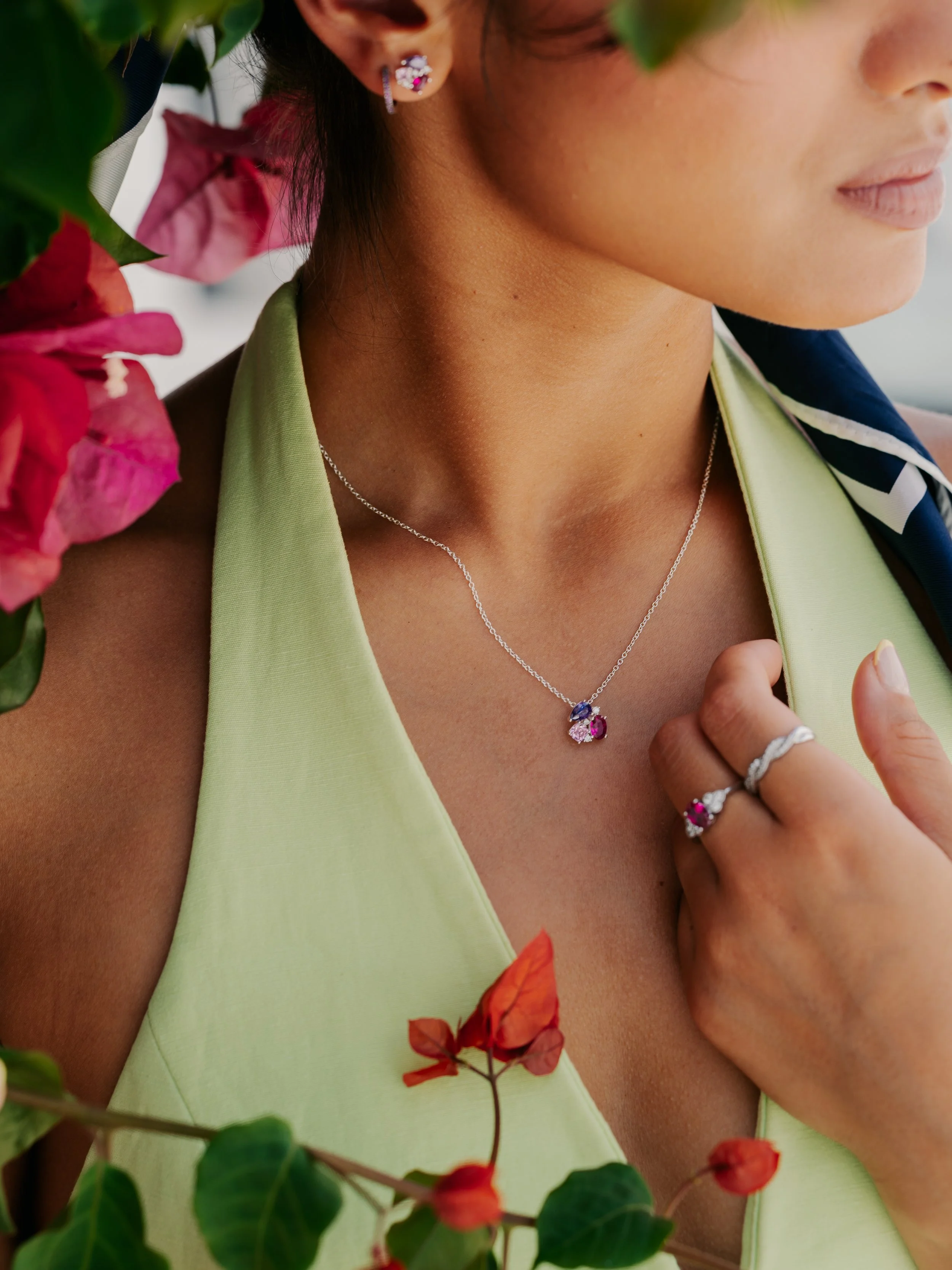 Close-up of a woman wearing a necklace with a pink and purple gemstone pendant, earrings, and rings, surrounded by pink and red flowers.