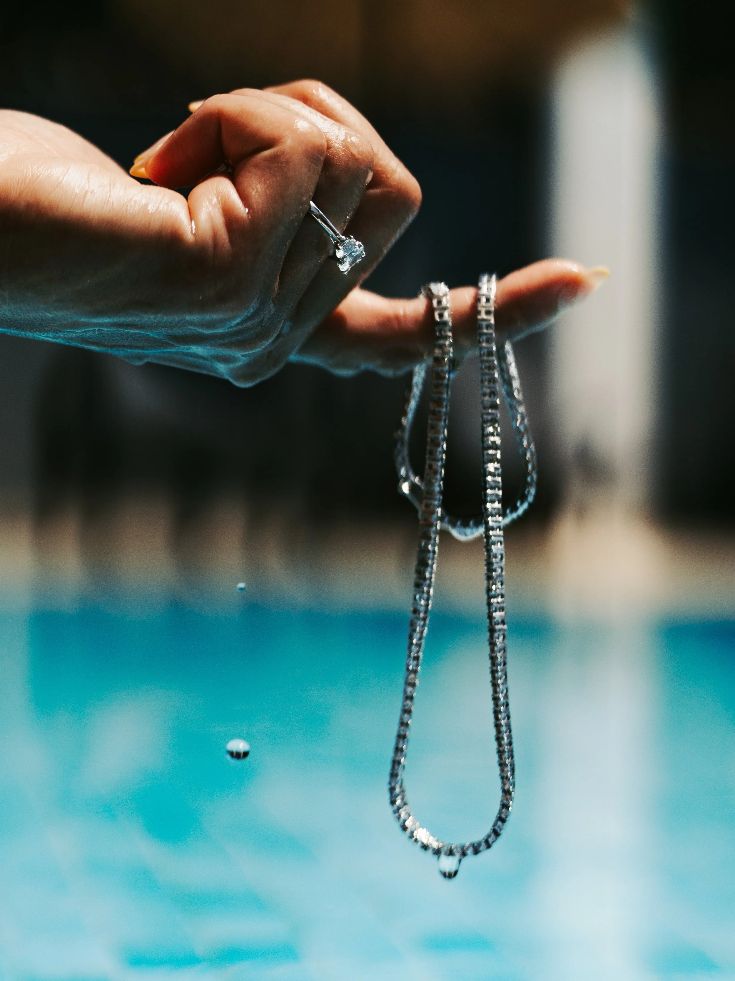 Close-up of a hand holding a diamond ring and a necklace, with water droplets falling.