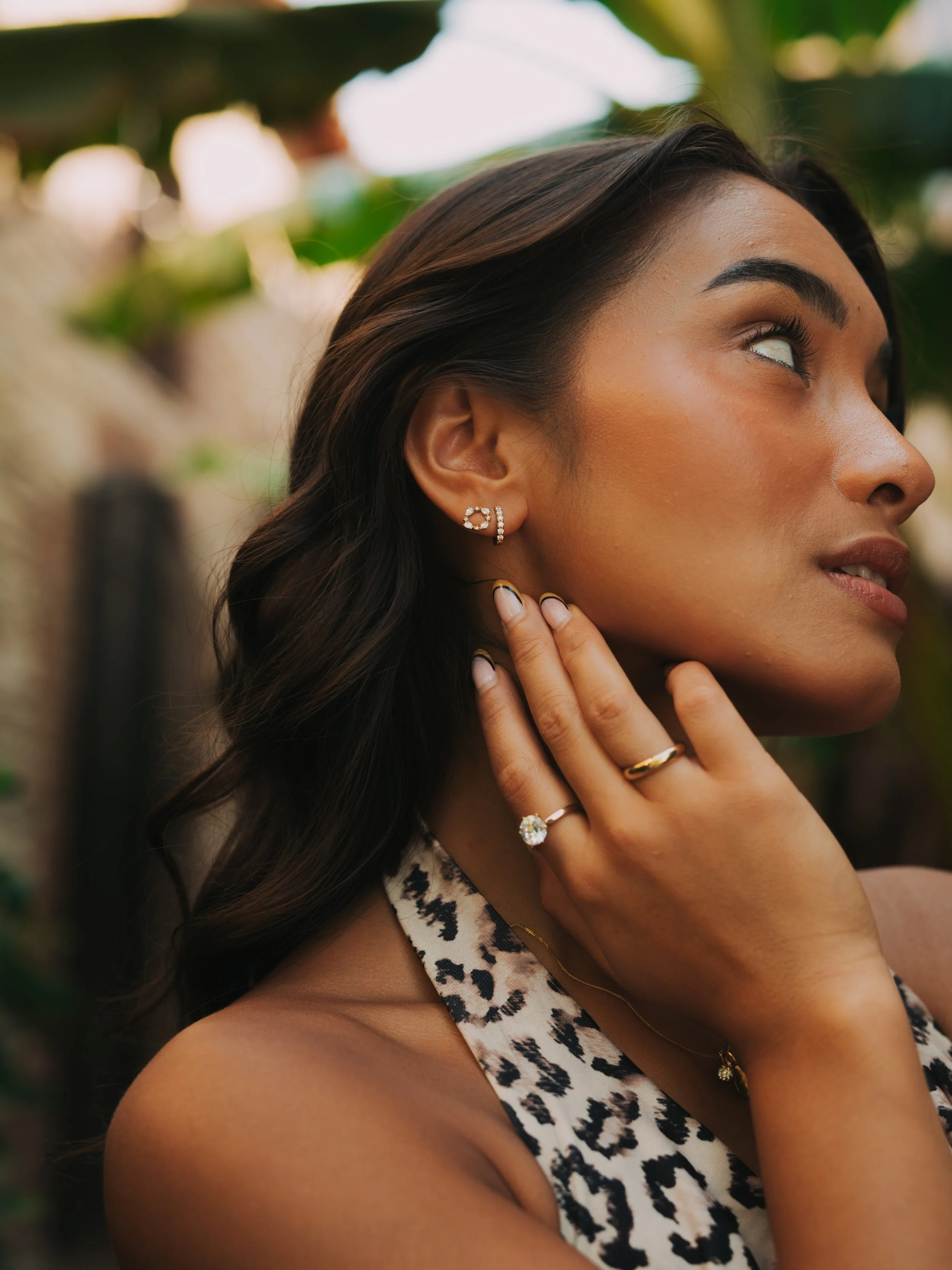 Close-up of a woman with dark brown hair, wearing jewelry including earrings, rings, and a necklace, in an outdoor setting with greenery in the background.