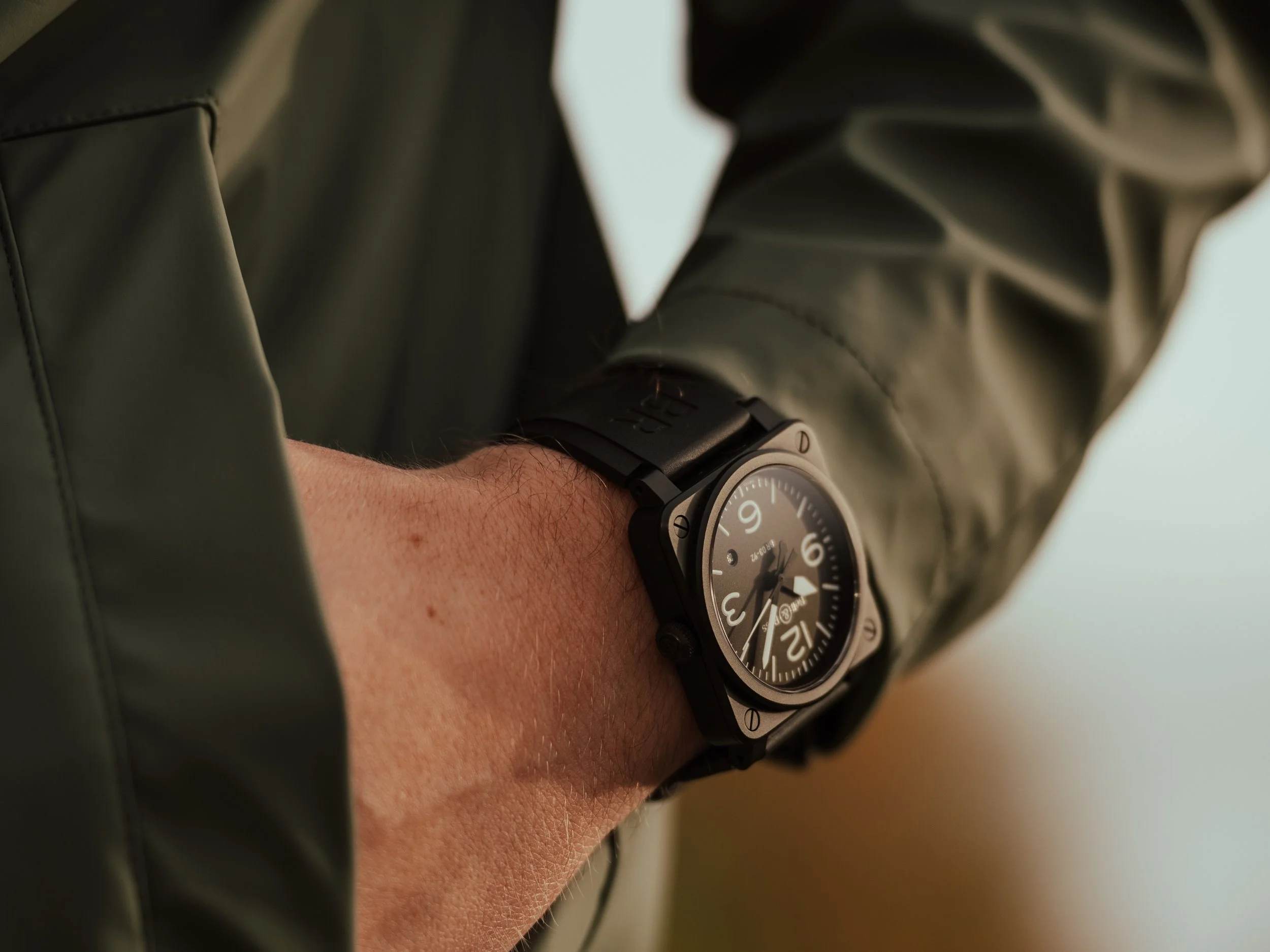 A close-up of a person's wrist wearing a black wristwatch with a round face, black band, and black dial showing white numbers, leather jacket sleeve nearby.