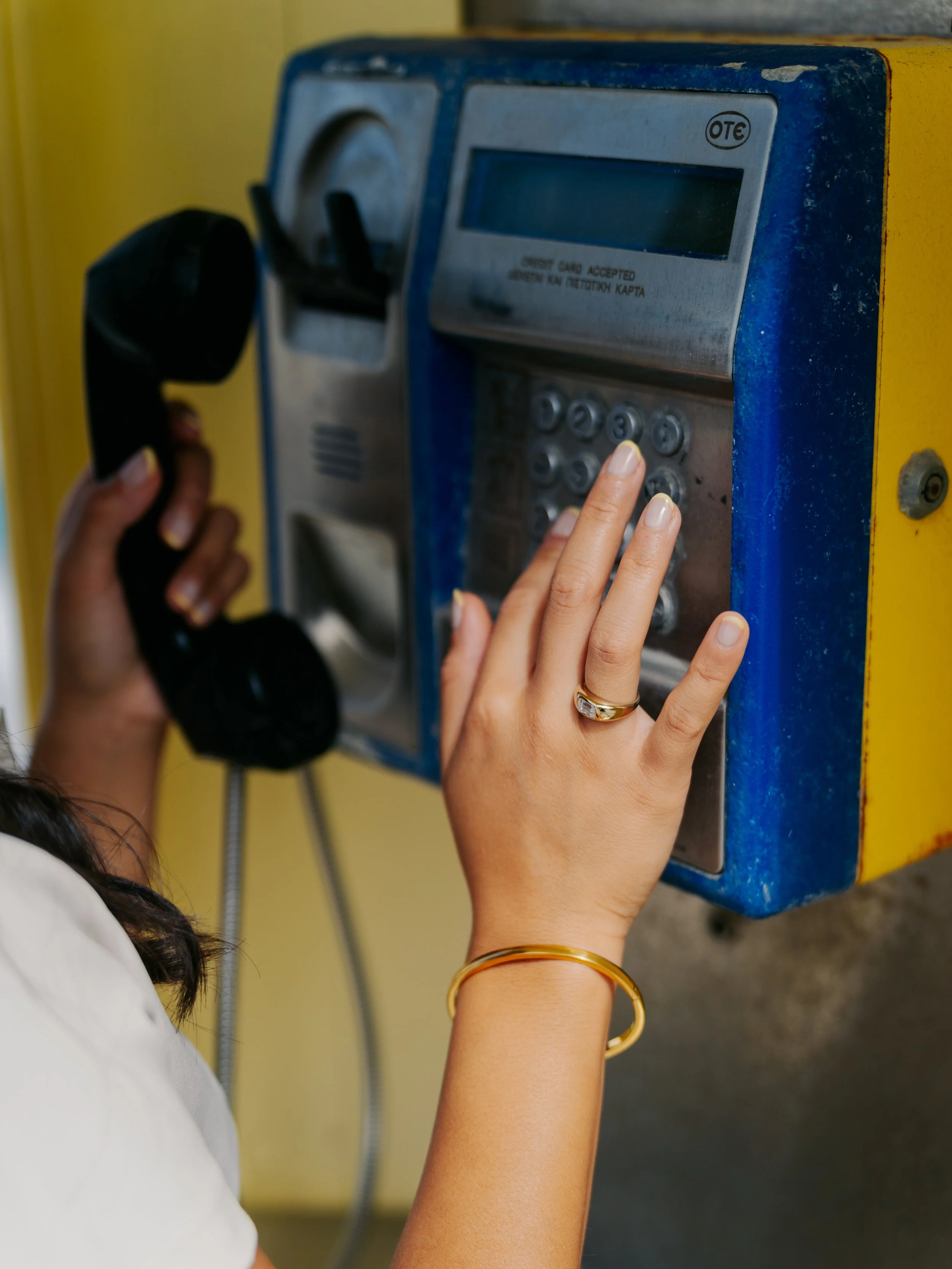 Person about to press keypad on public payphone, holding the receiver with one hand and wearing a gold ring and bracelet.