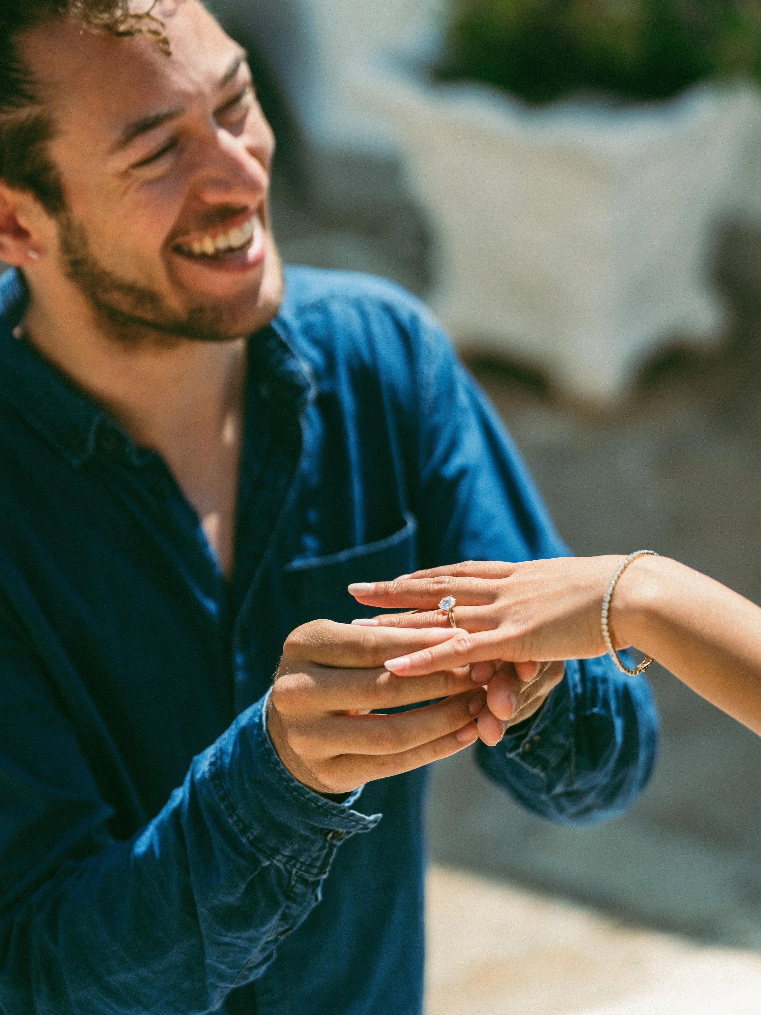 A man is smiling and holding a woman's hand, which has a large engagement ring, during a proposal at a scenic outdoor location.