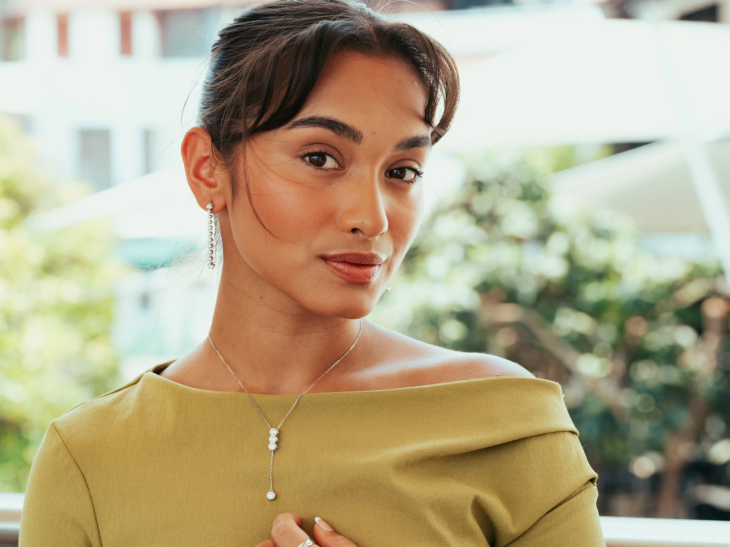 A woman with dark hair styled back, wearing earrings and a matching necklace, dressed in a yellow off-the-shoulder top, with a blurred outdoor background of trees and buildings.