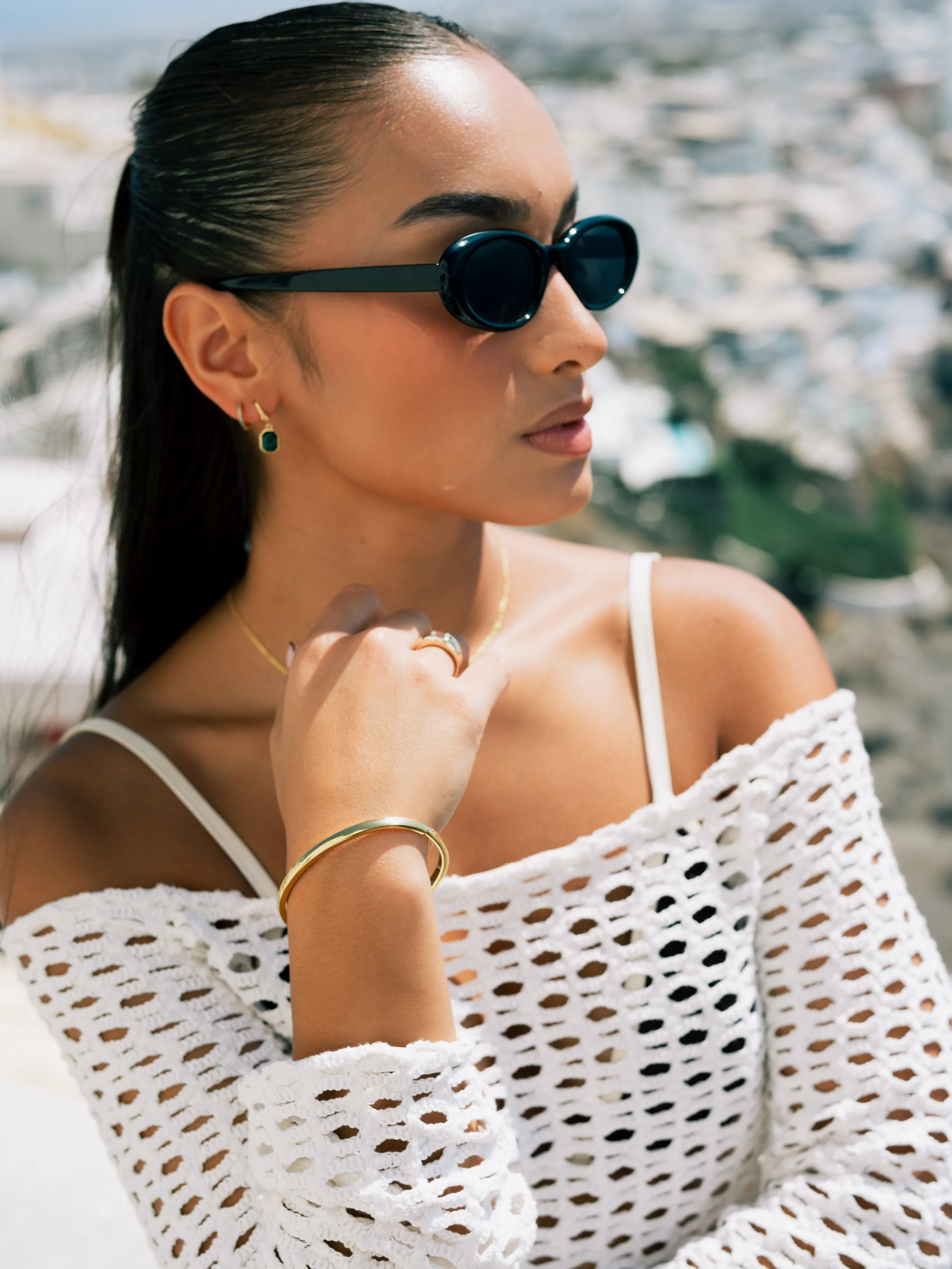 A woman wearing dark sunglasses, gold jewelry, and a white crocheted off-shoulder top outdoors with a blurred cityscape in the background.