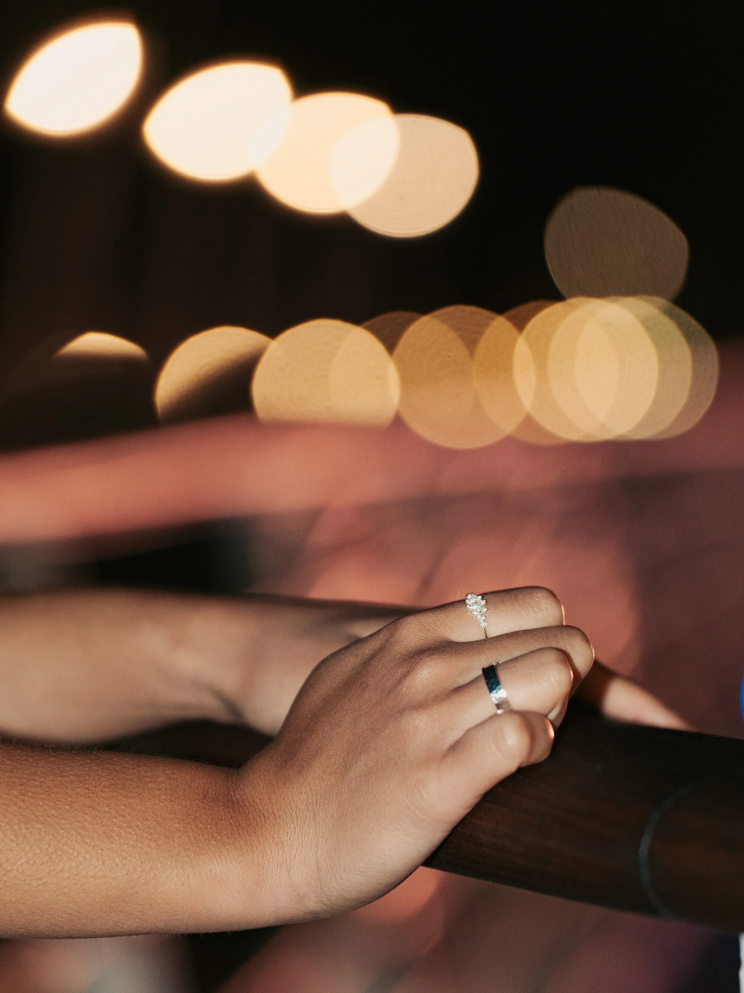 Close-up of a person's left hand with two rings resting on a railing at night, with blurred city lights in the background.