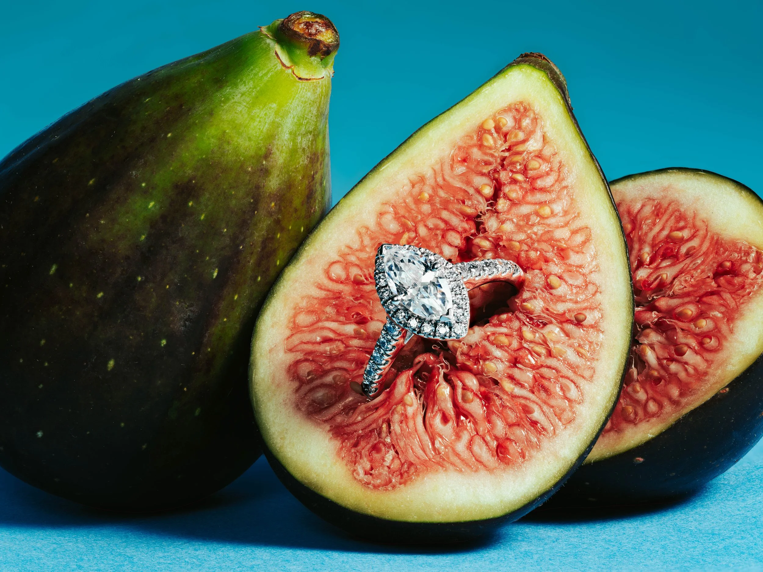A green fig fruit cut in half with a diamond engagement ring placed inside the cut, against a blue background.