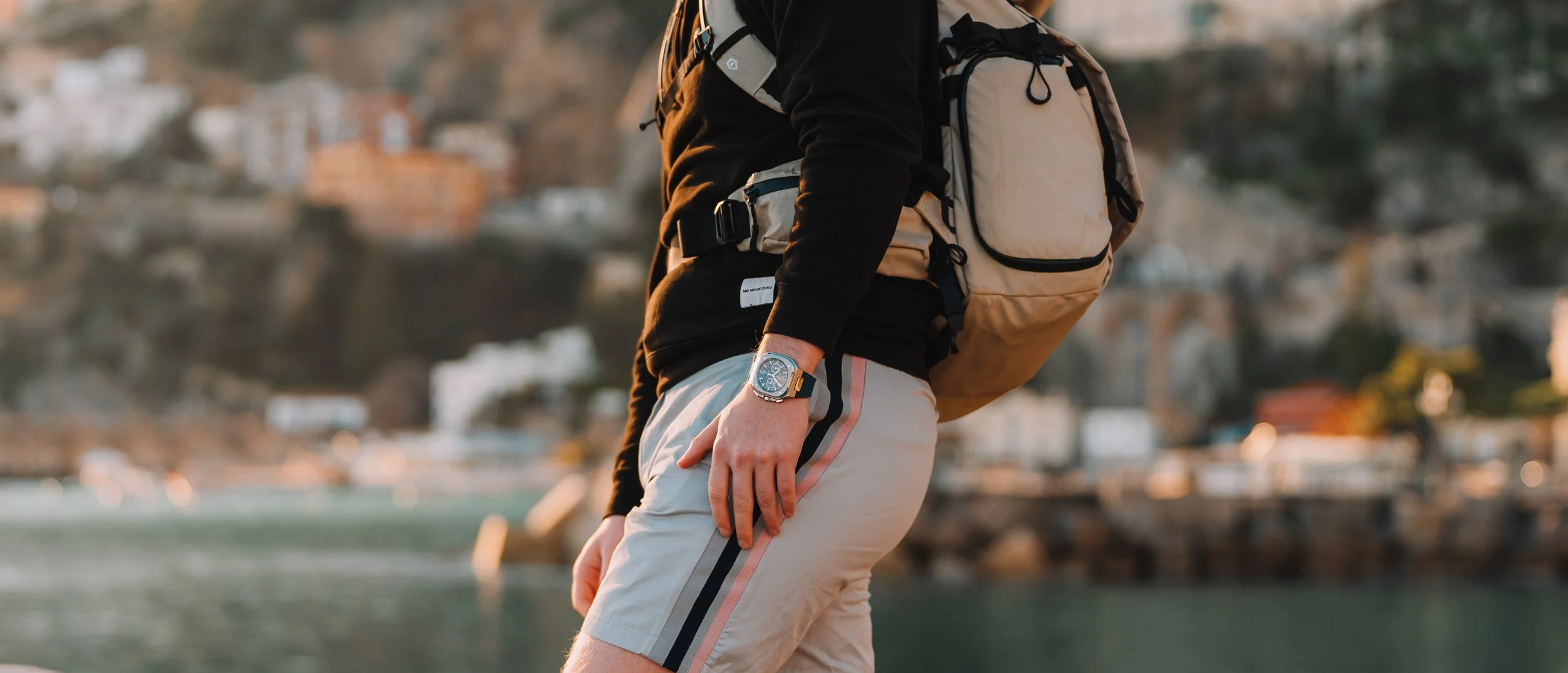 Close-up of a hiker wearing a black long-sleeve shirt, beige shorts with a stripe, a smartwatch, and carrying a beige backpack by a waterfront with houses and hills in the background.
