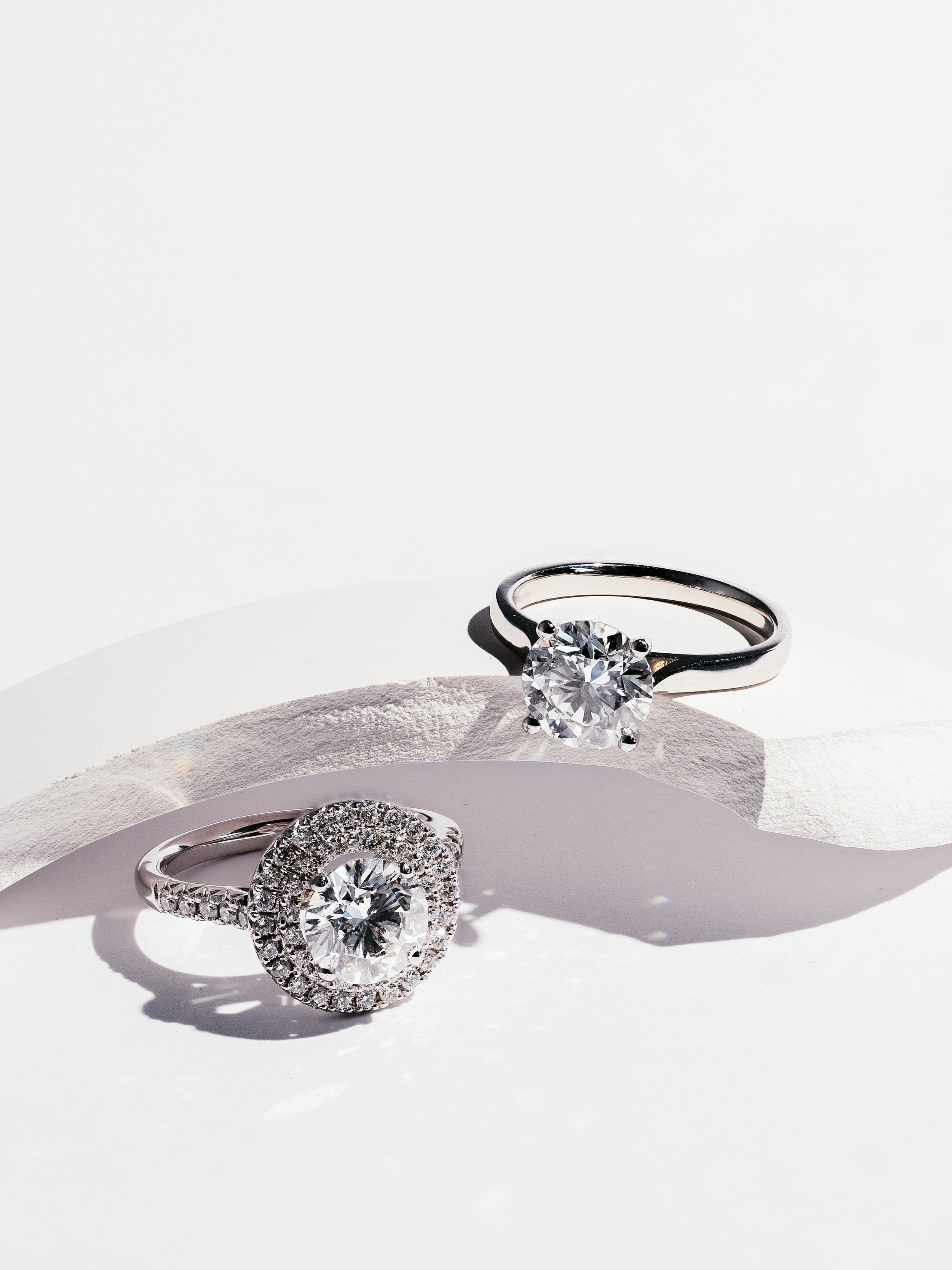 Two silver rings with large diamonds, one with a round center stone surrounded by smaller diamonds, displayed on white surface with shadows.