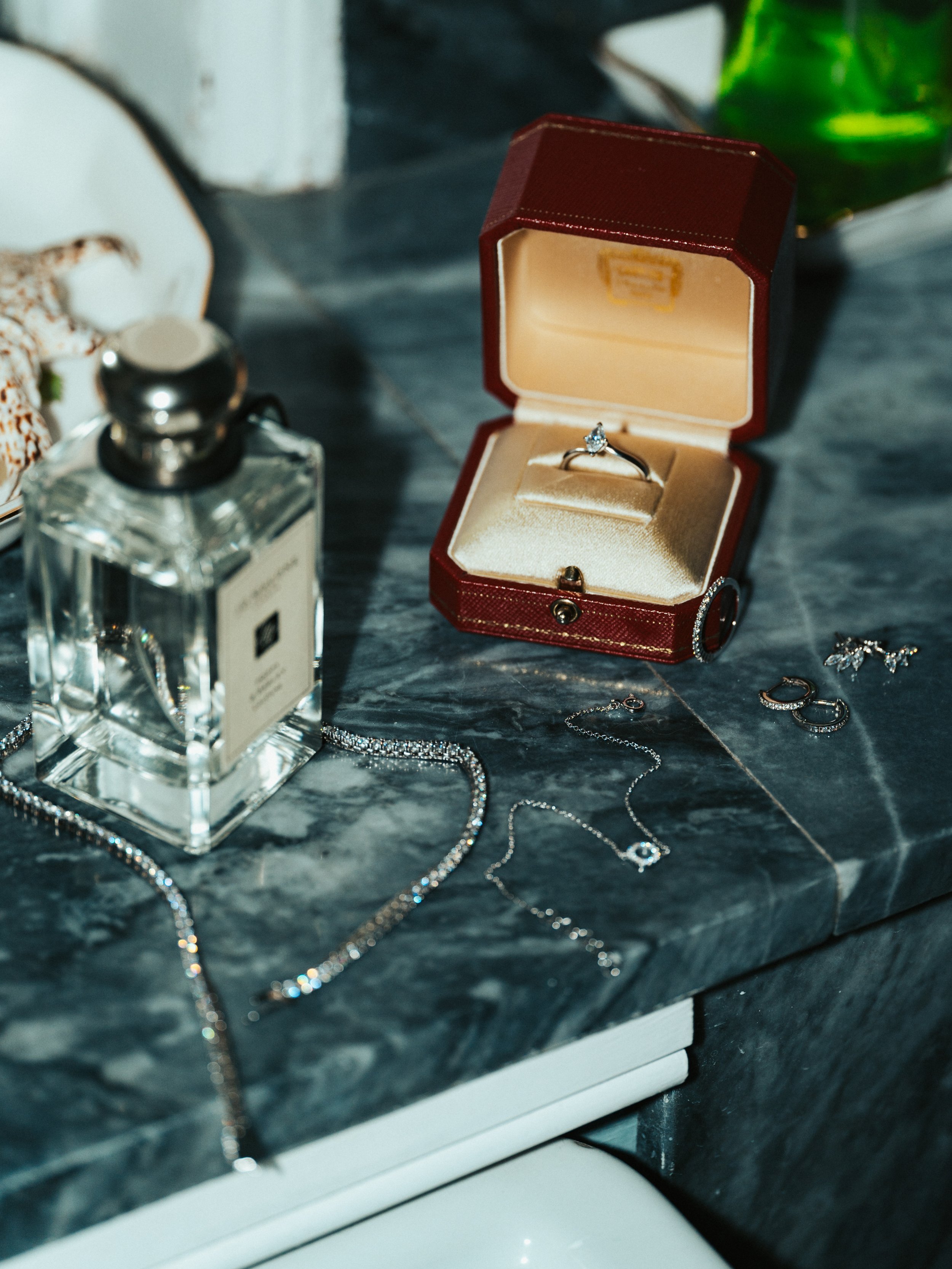 A jewelry collection on a marble surface, including a diamond engagement ring in a red box, various necklaces, earrings, and a bottle of perfume.