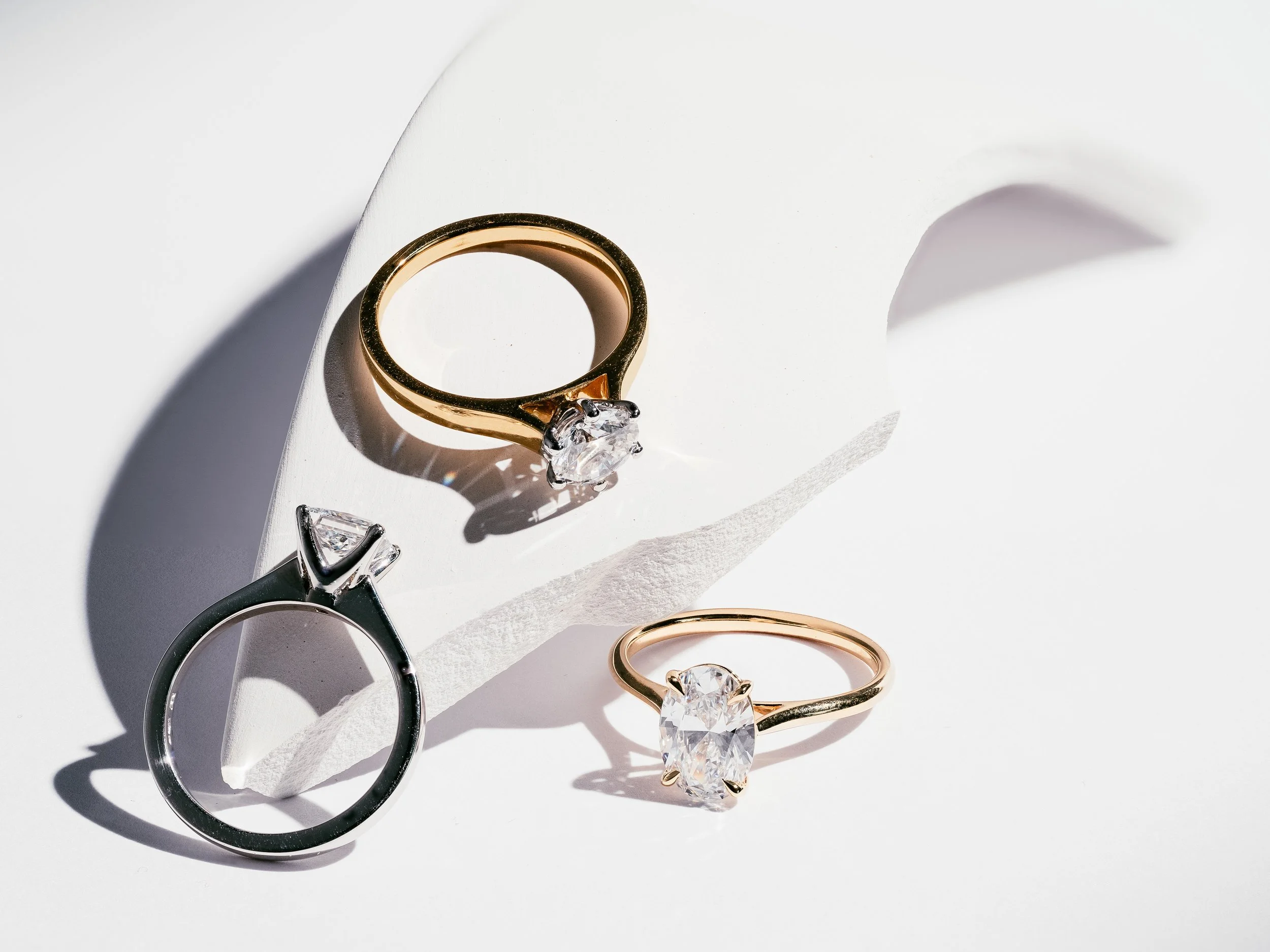 Three rings with large central gemstones placed on a white curved surface with shadows.