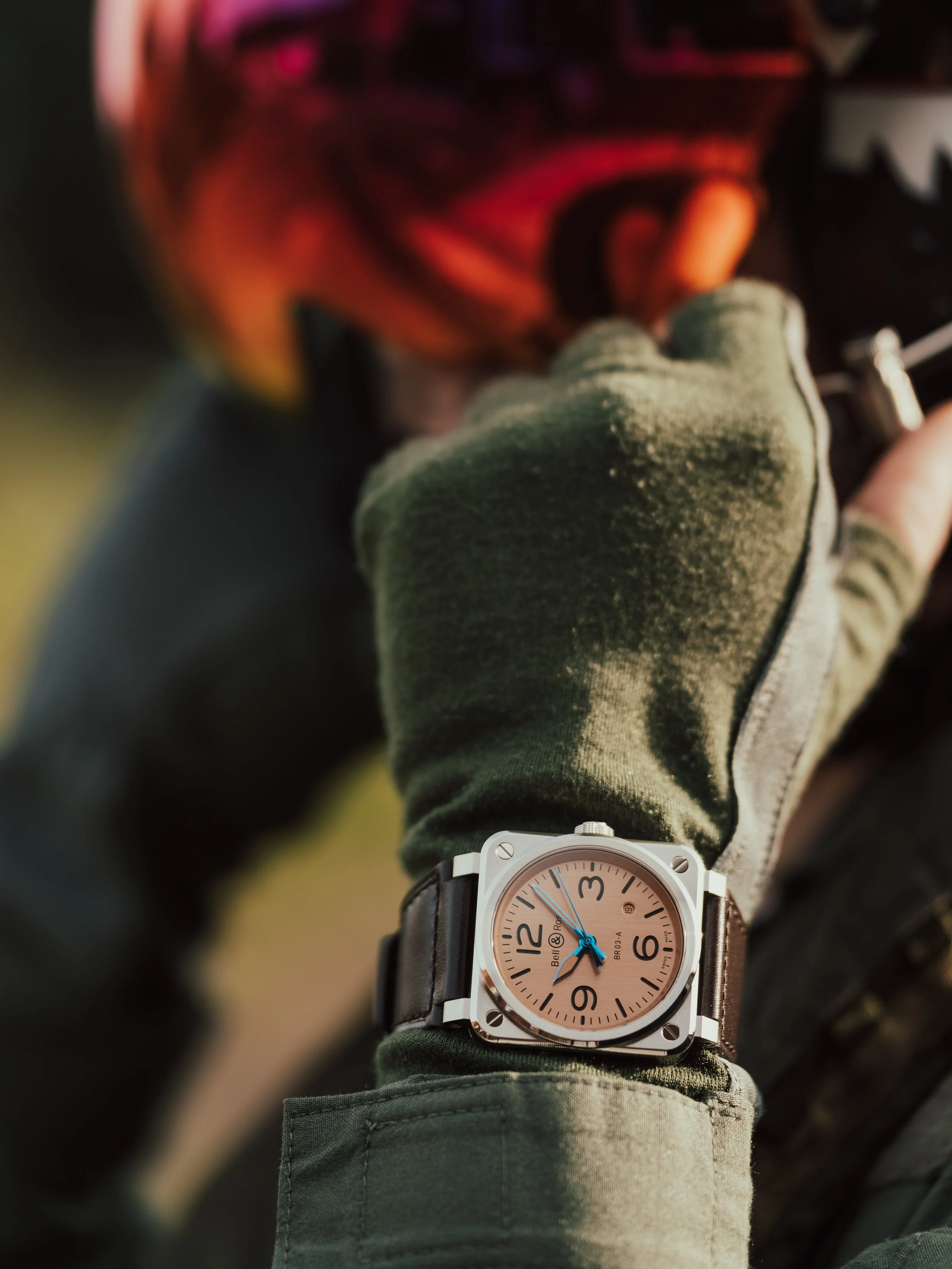 Close-up of a wristwatch with a beige face and black leather strap on a person's wrist, wearing a green jacket and green glove, outdoors with a blurred background.