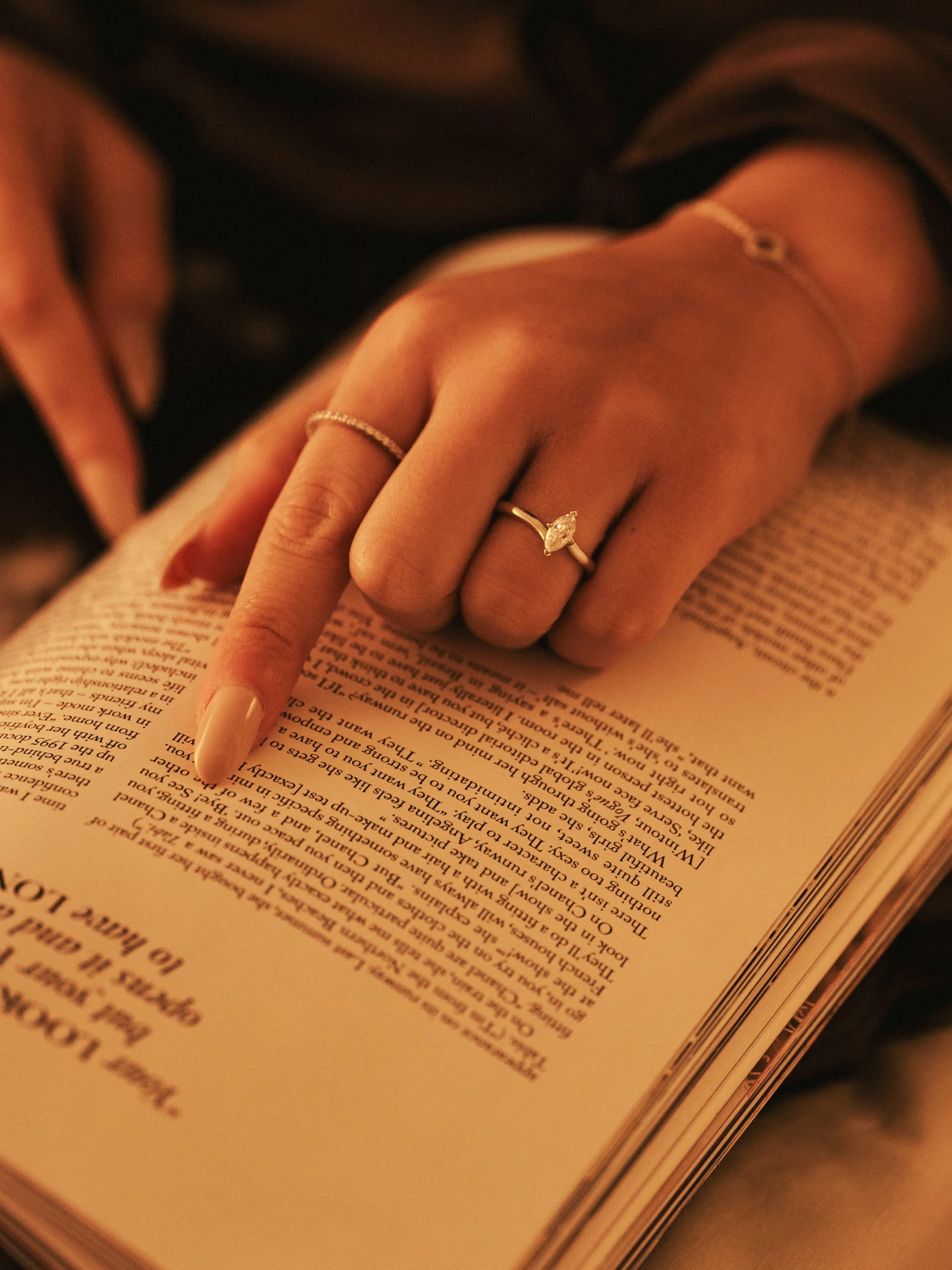 A person's hand with rings resting on an open book, with one finger pointing to a paragraph.