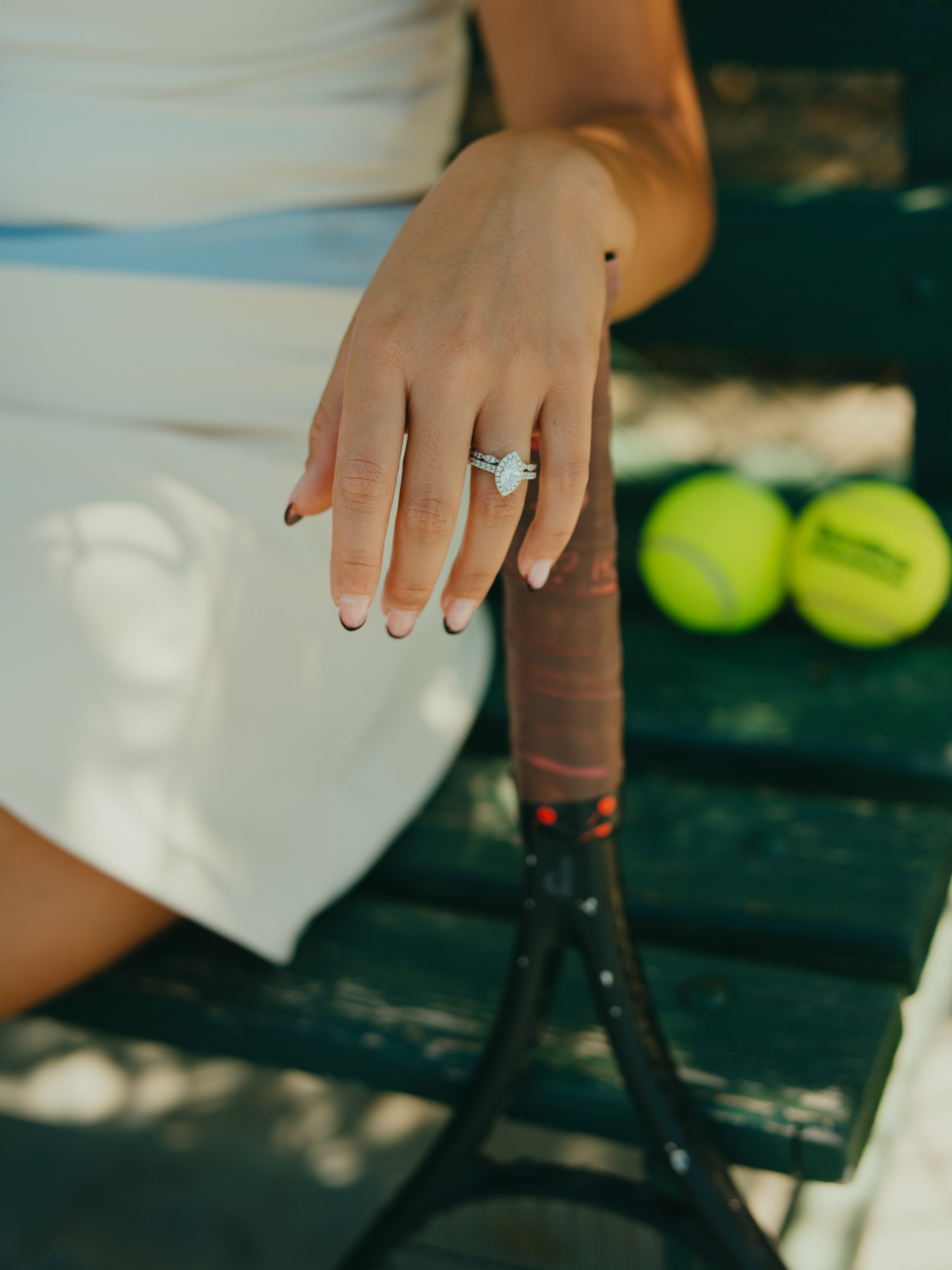 Close-up of a woman's hand with an engagement ring resting on a tennis racket, with tennis balls on a nearby bench.