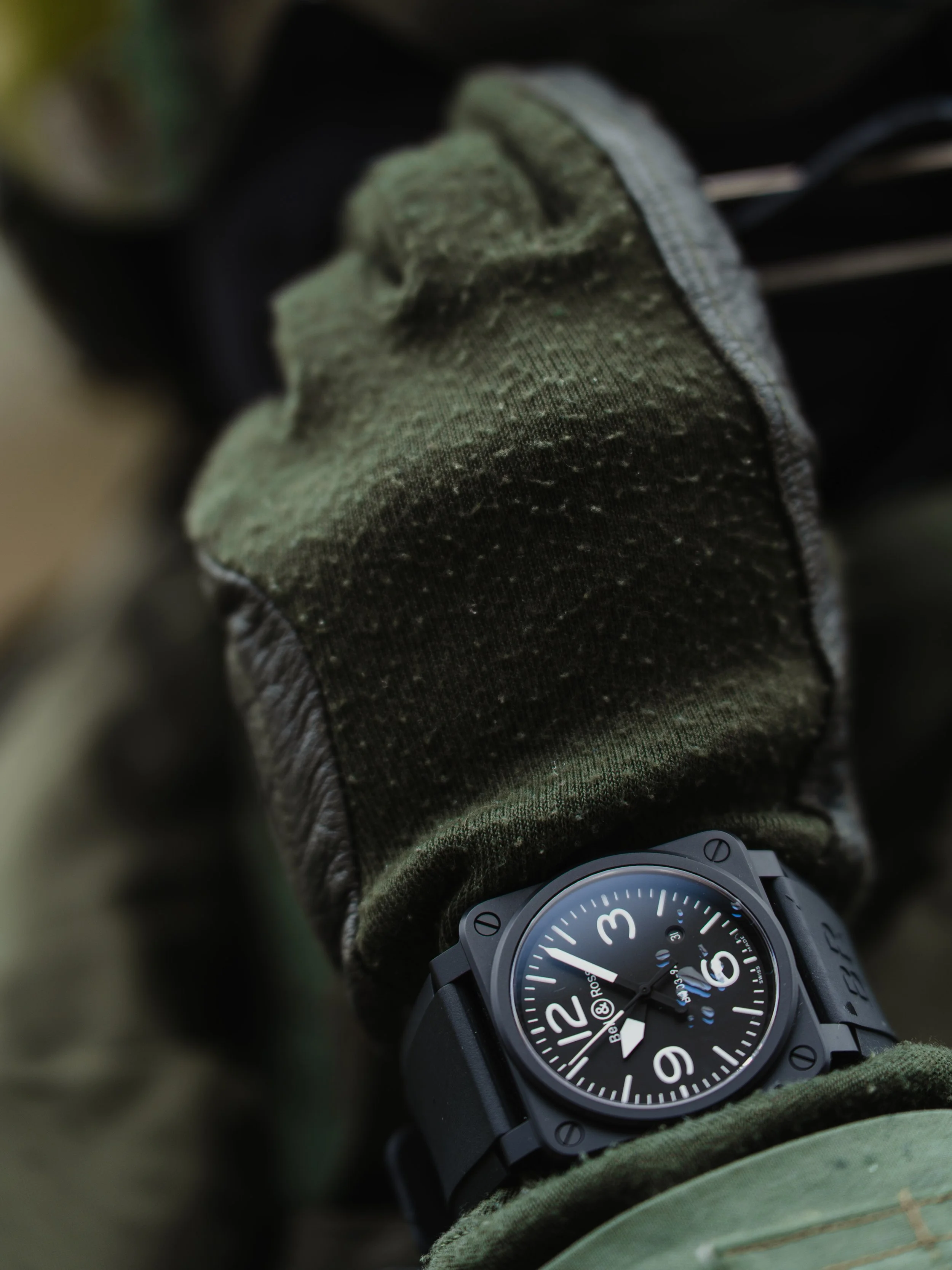 Close-up of a black wristwatch with a large round dial on a person's wrist, partially covered by a green glove or sleeve.