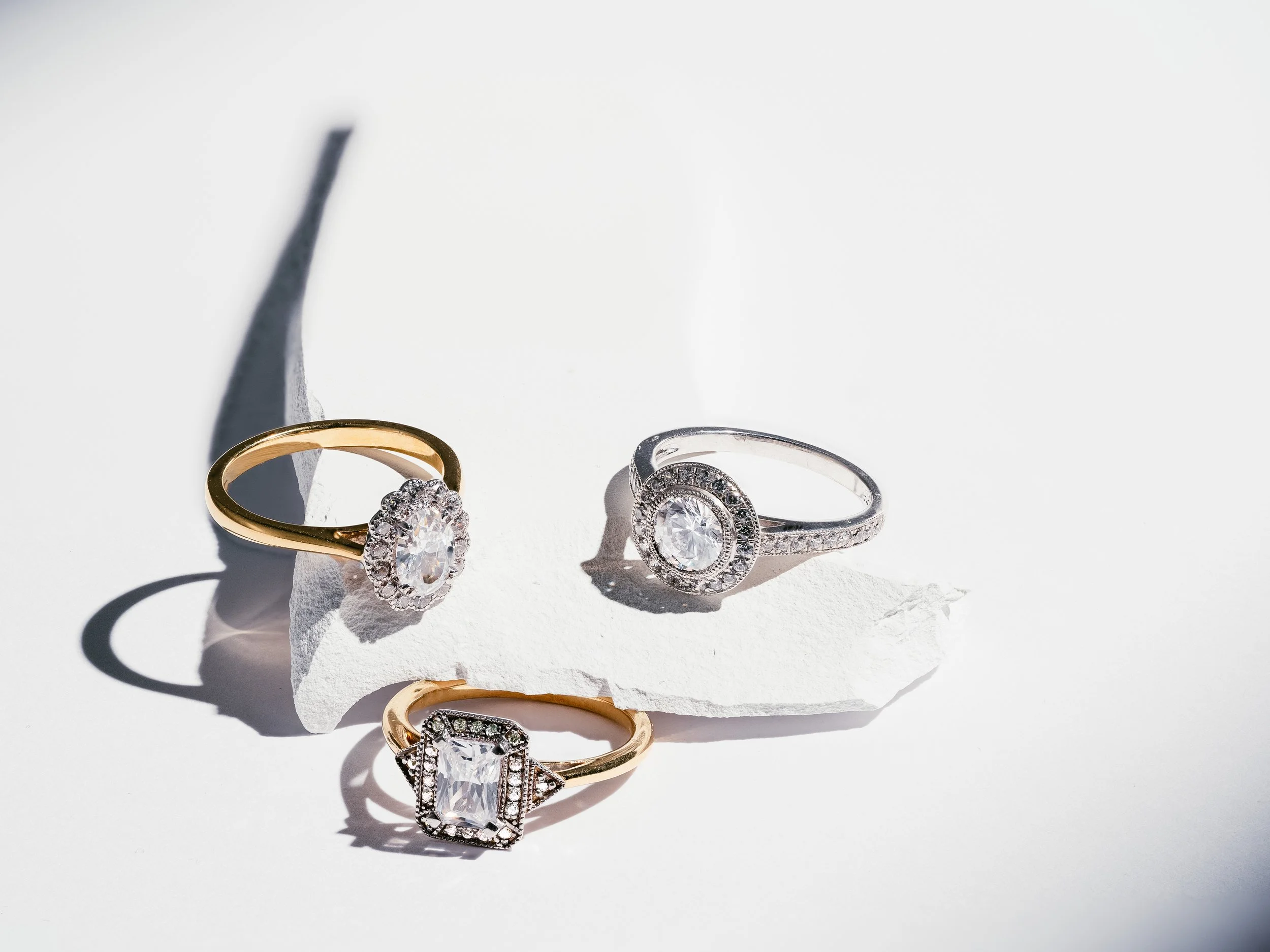 Three rings with large central stones surrounded by smaller stones, displayed on a white surface with shadows