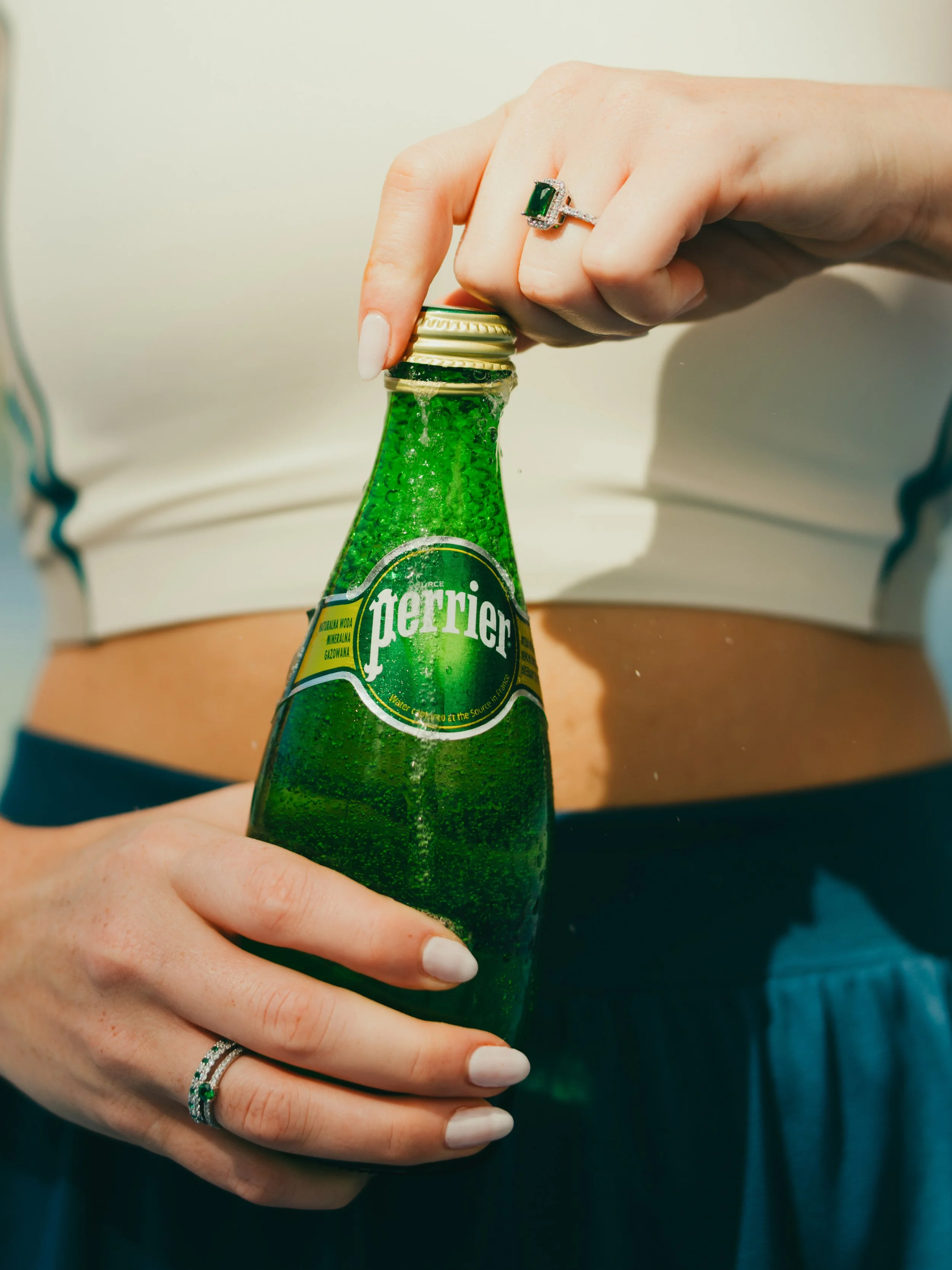 Person holding a green Perrier sparkling water bottle with a ring on their finger, standing outdoors in sunlight.