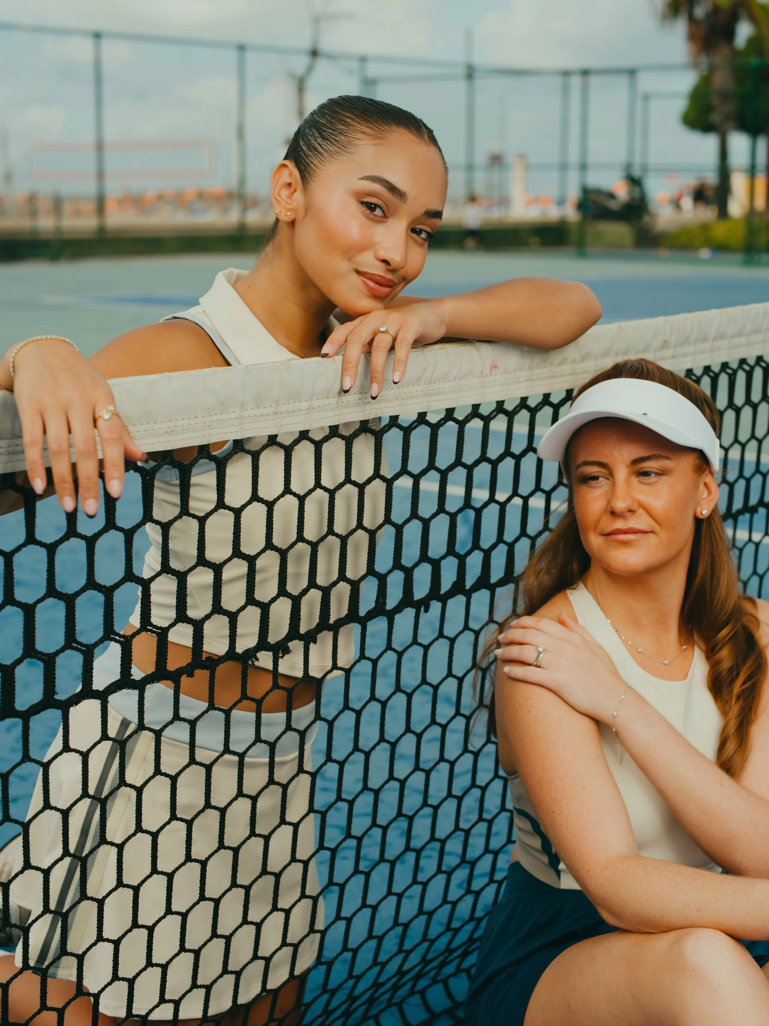 Two women at a tennis court, one leaning on the net and the other sitting beside it, both dressed in tennis outfits with one wearing a visor.