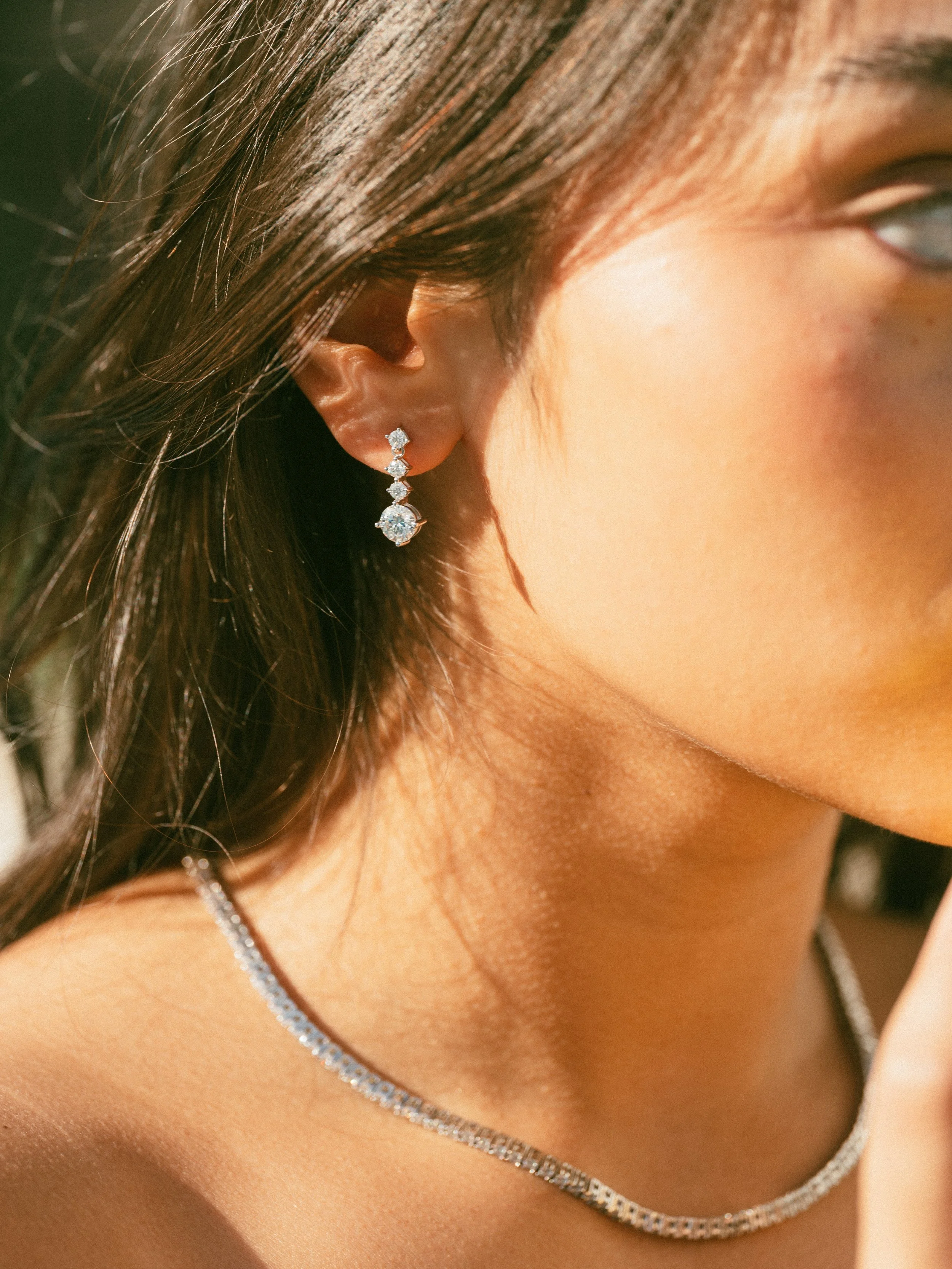 Close-up of a woman's face and shoulder, showcasing diamond earrings and a diamond necklace.