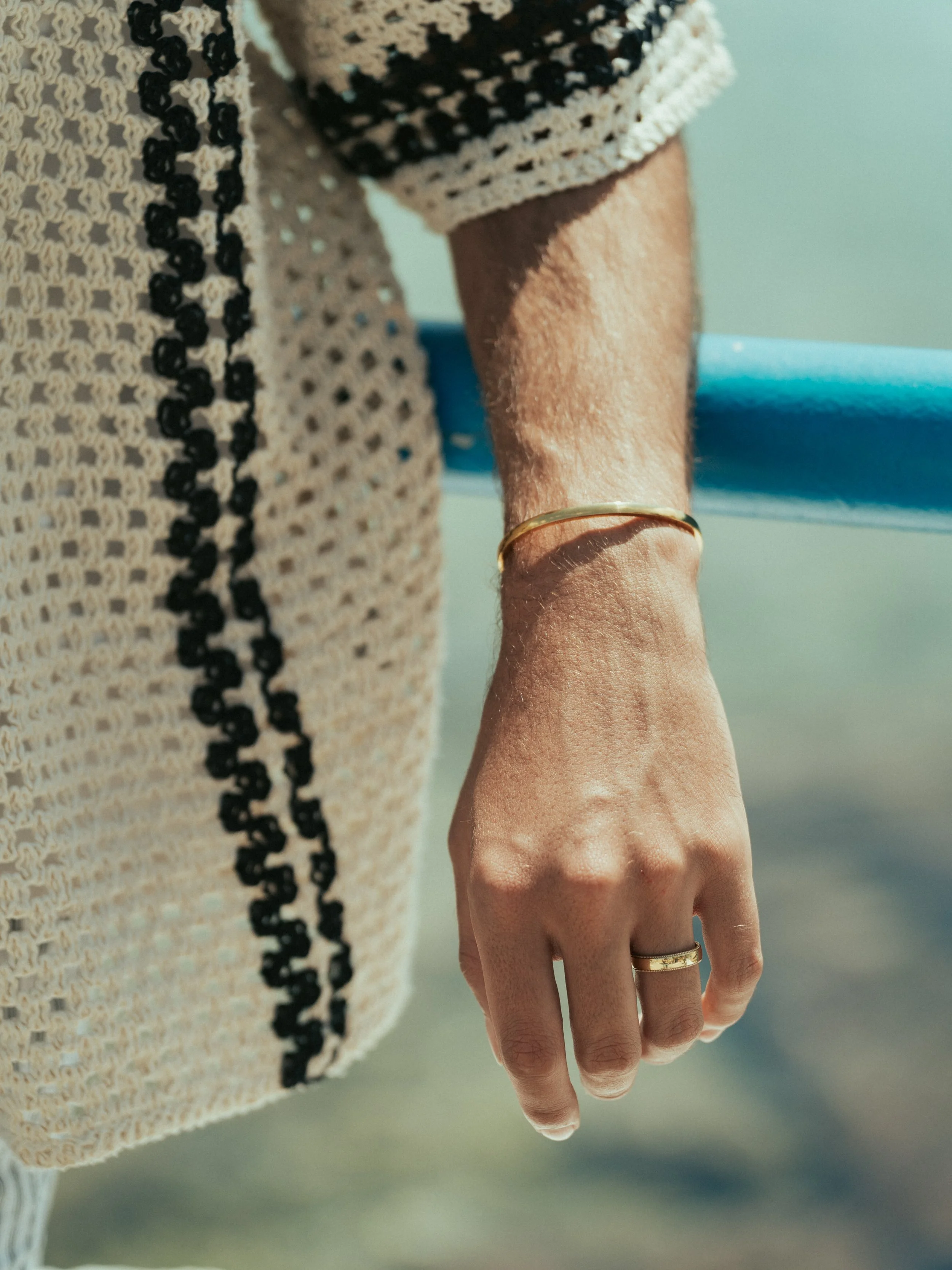 Close-up of a person's hand with rings and a gold bracelet, holding onto a blue bar on a boat or dock, with a sandy beach in the background.