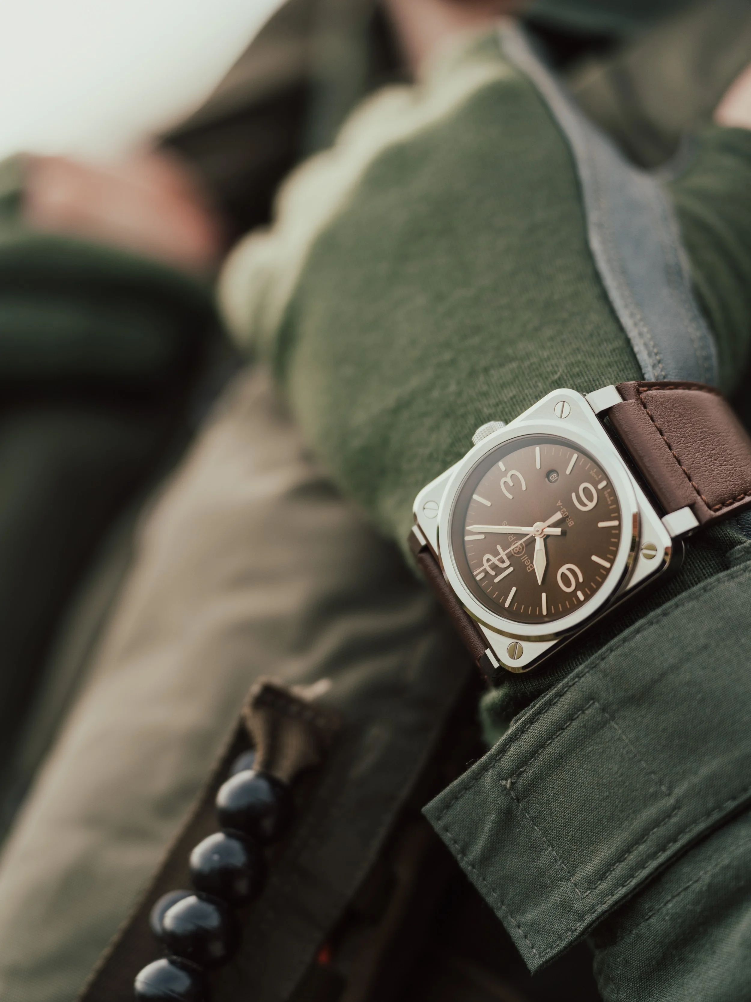 A wristwatch with a brown leather strap worn on a person's wrist, showing a black dial with white numerals and hands, resting on a green jacket.