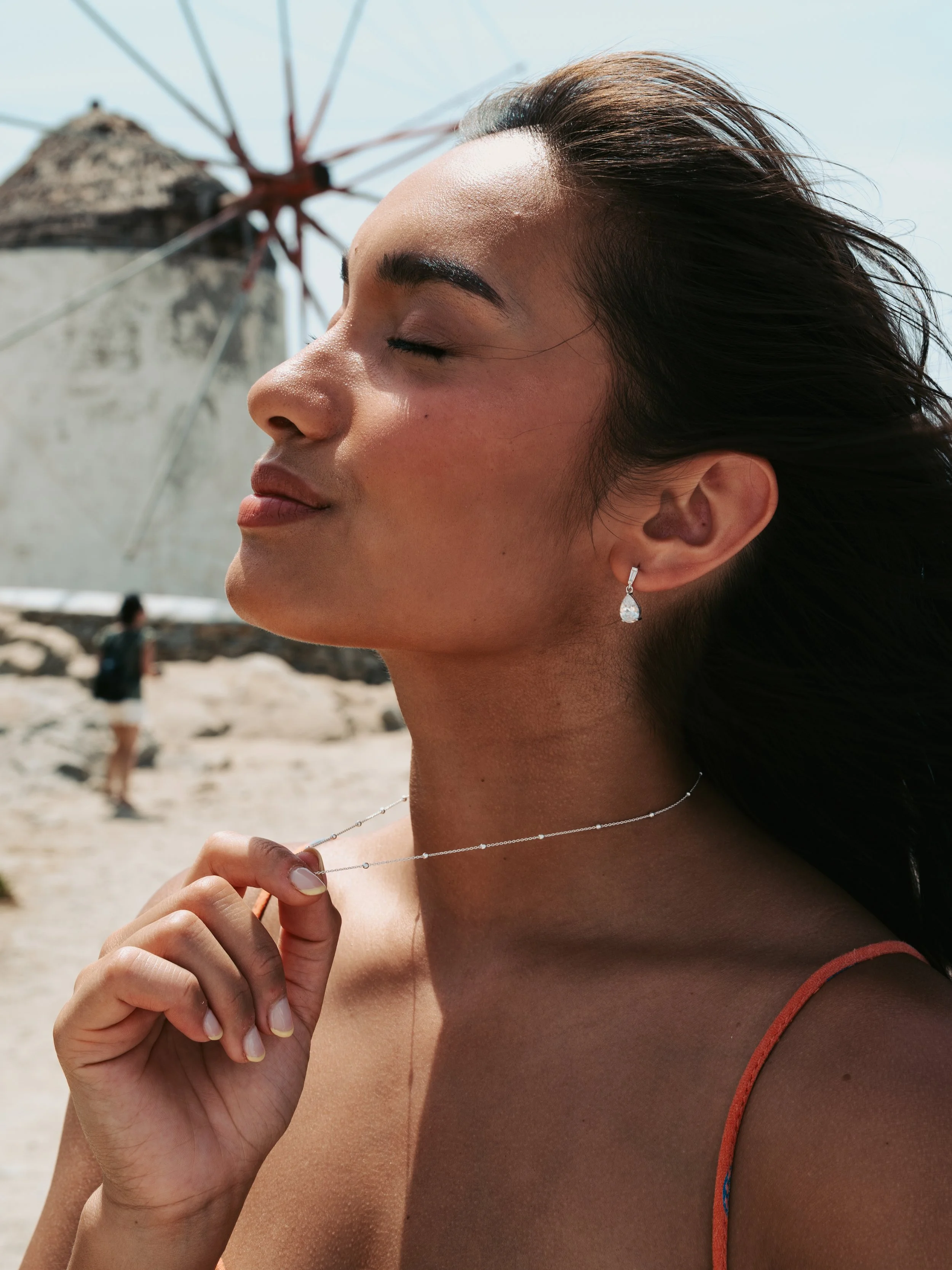 A woman with long dark hair, wearing earrings and a necklace, stands on a beach with her eyes closed, enjoying the sun, with a beach umbrella and another person in the background.
