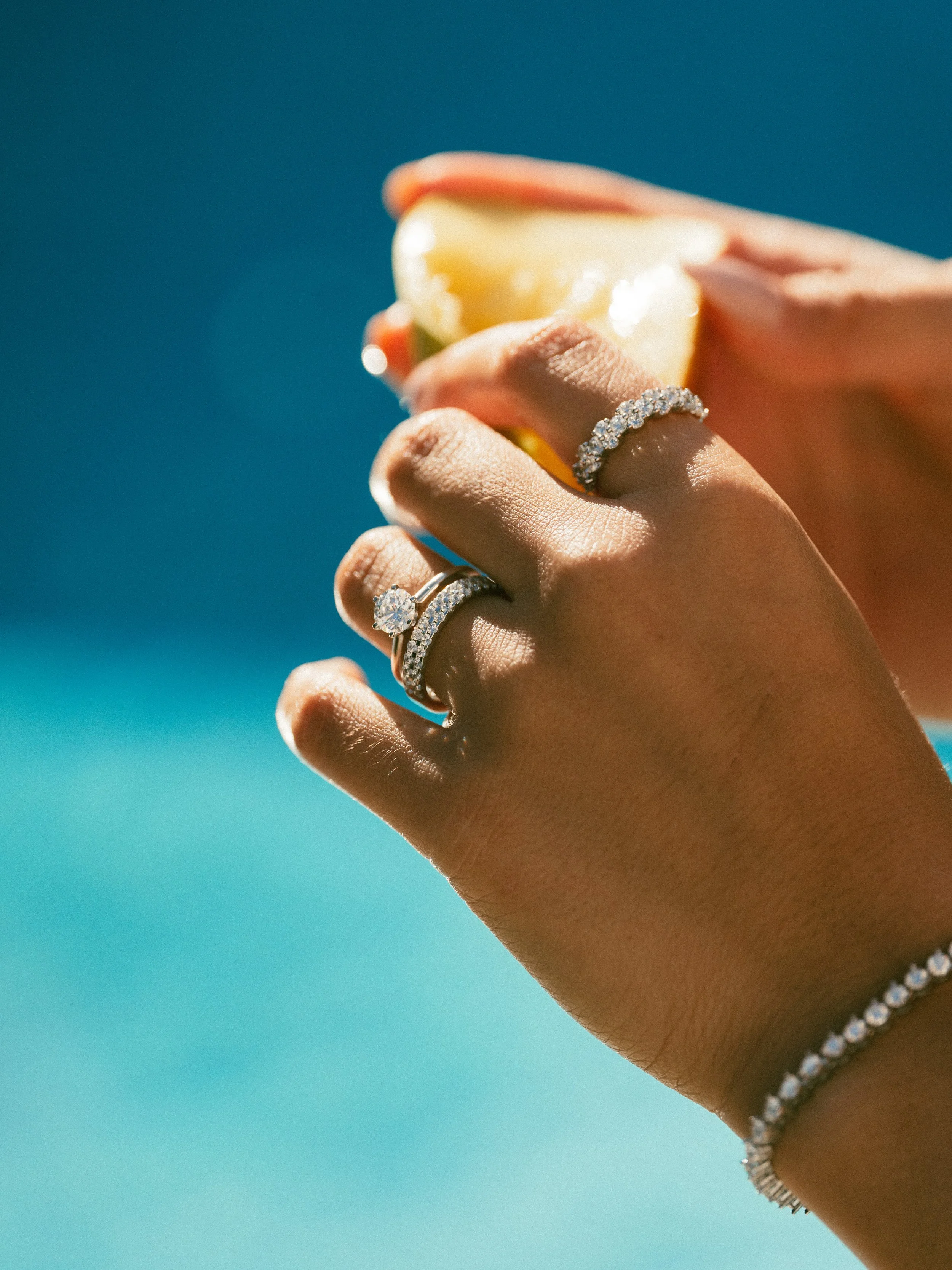 Close-up of a hand wearing diamond rings and a bracelet, holding a lemon wedge with a blue background.